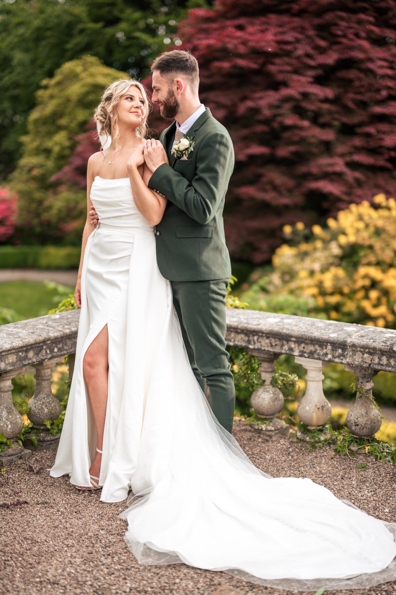 A bride in a white gown and groom in a green suit embrace in a lush garden setting on their wedding day.