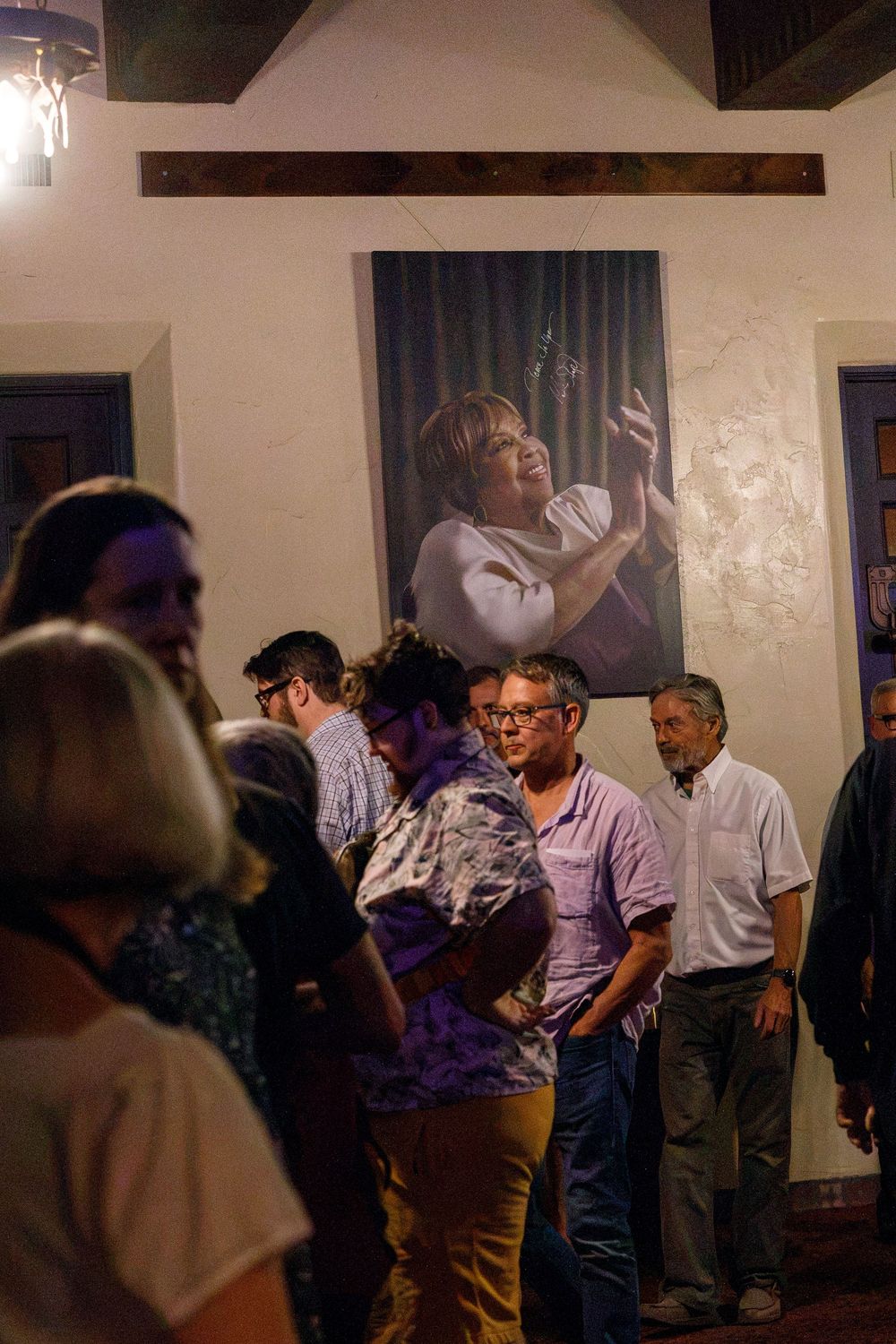 Guests mingle at an indoor gallery event with a large portrait of a woman singing displayed on the wall.