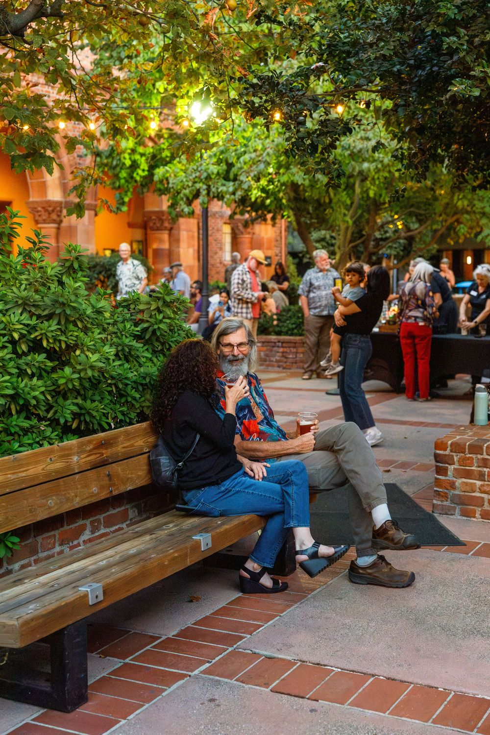 Couple sitting on wooden bench in courtyard garden at evening social gathering with guests mingling nearby.