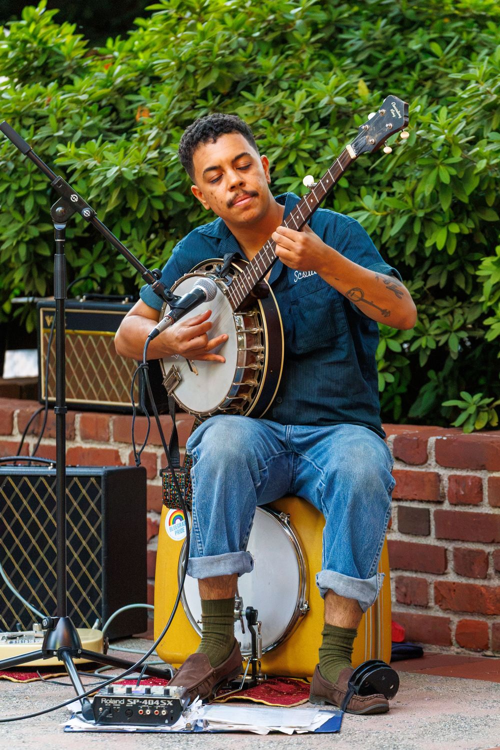 A musician plays banjo while seated outdoors against a brick wall with green foliage backdrop.