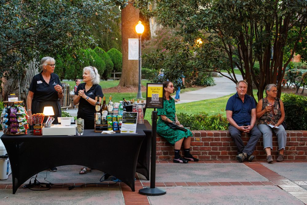 Outdoor evening event with speakers at a table and seated guests on a brick patio surrounded by trees.