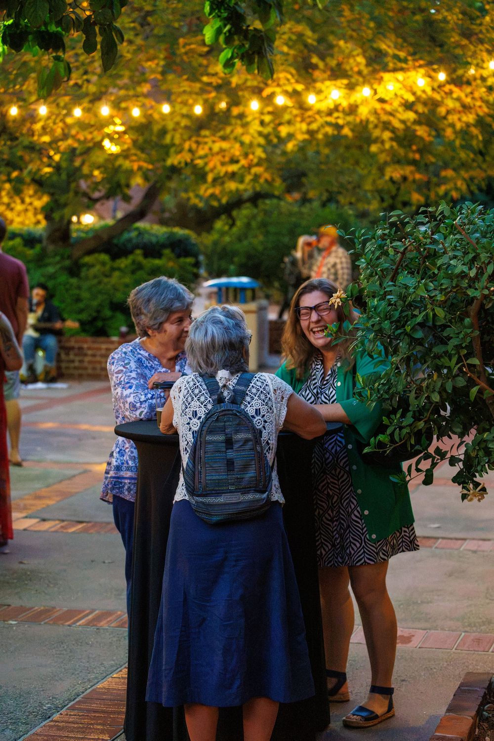Three women laughing and conversing outdoors at an evening garden gathering with warm string lights glowing above.