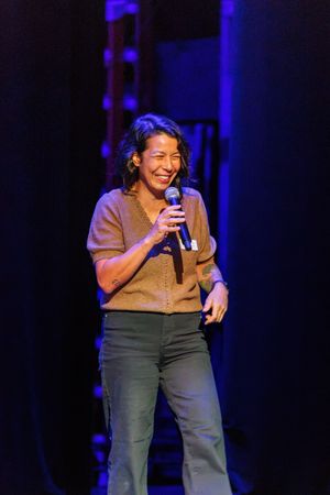 Woman smiling on stage holding a microphone under blue stage lights during a live performance.