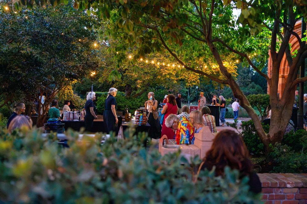 Guests dine outdoors under string lights surrounded by lush trees at a garden evening event.
