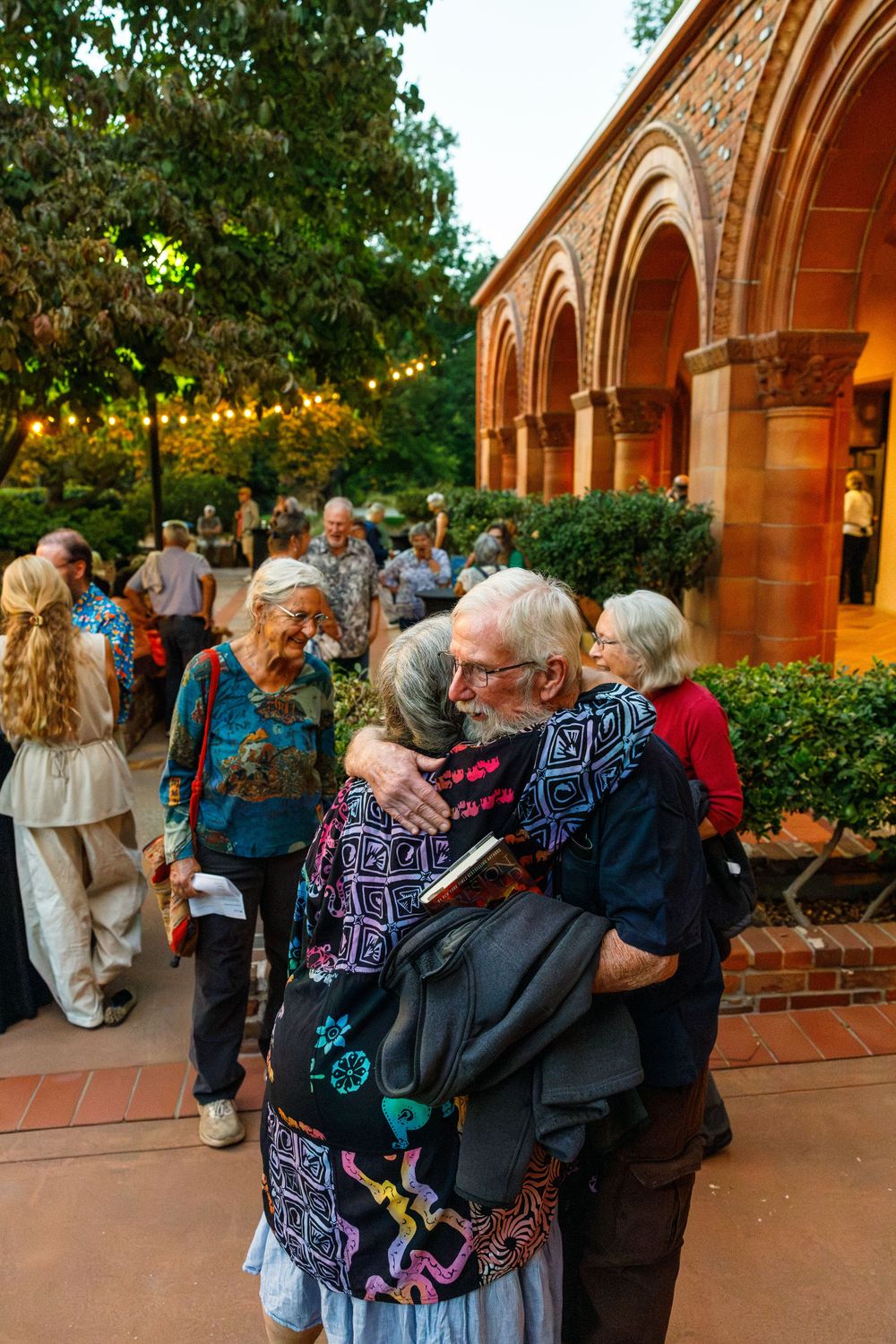 Elderly couple sharing a warm hug outdoors near a historic red brick building at a social gathering event.