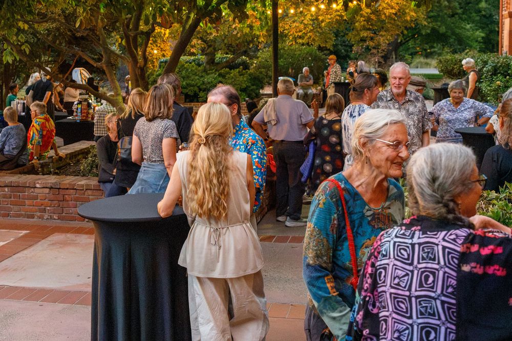 Outdoor evening garden party with diverse crowd of guests mingling on a brick patio surrounded by trees.