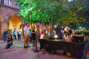 Guests gather at an outdoor evening event near a bar setup, surrounded by trees and historic brick architecture.