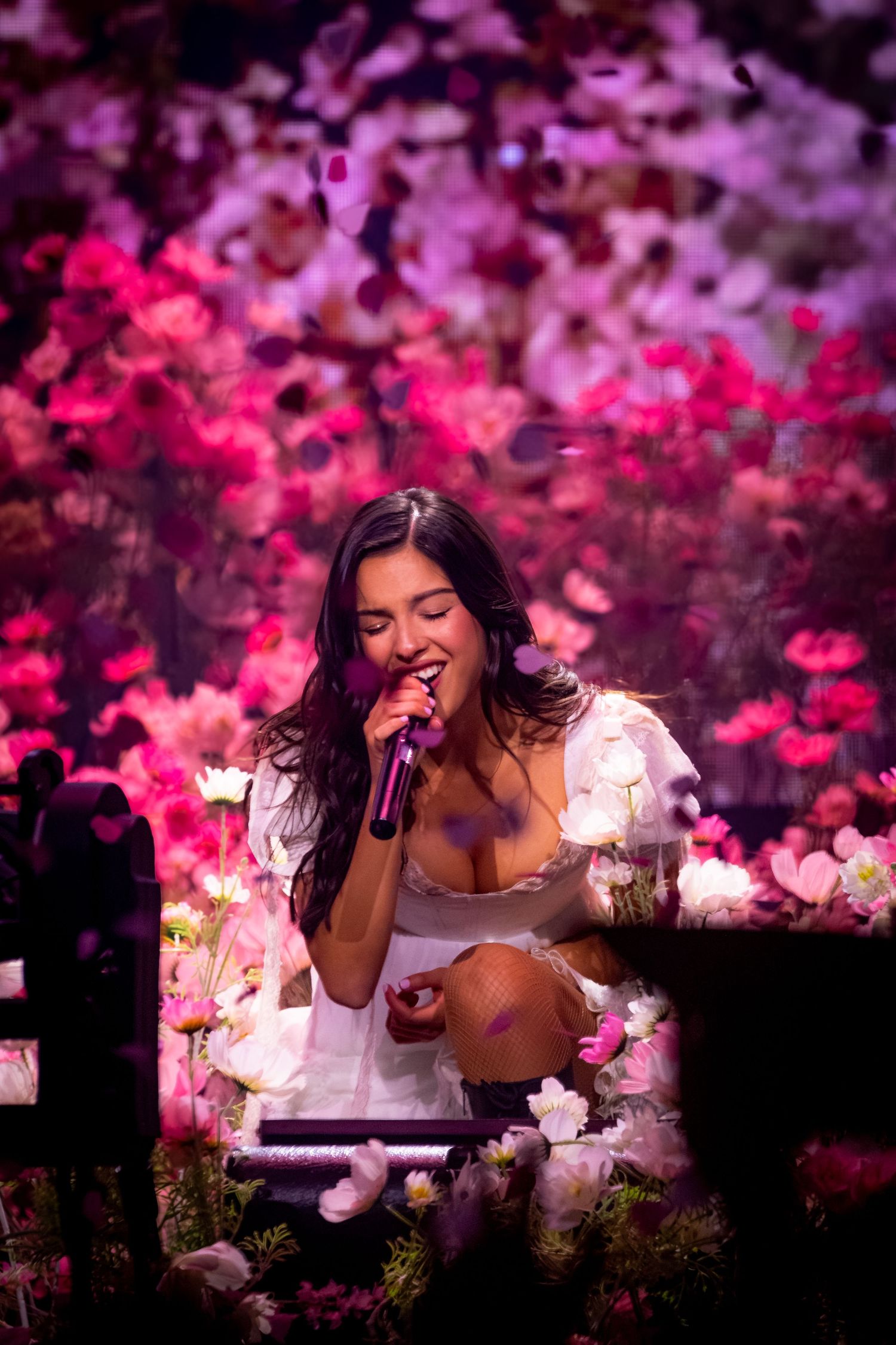 Olivia Rodrigo kneels and sings among pink and white flowers during opening night of the Unraveled Tour at PeoplesBank Arena in Hartford, CT