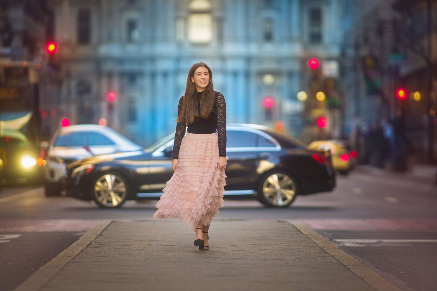 Elegant woman in pink tulle skirt walks across a city street at dusk.