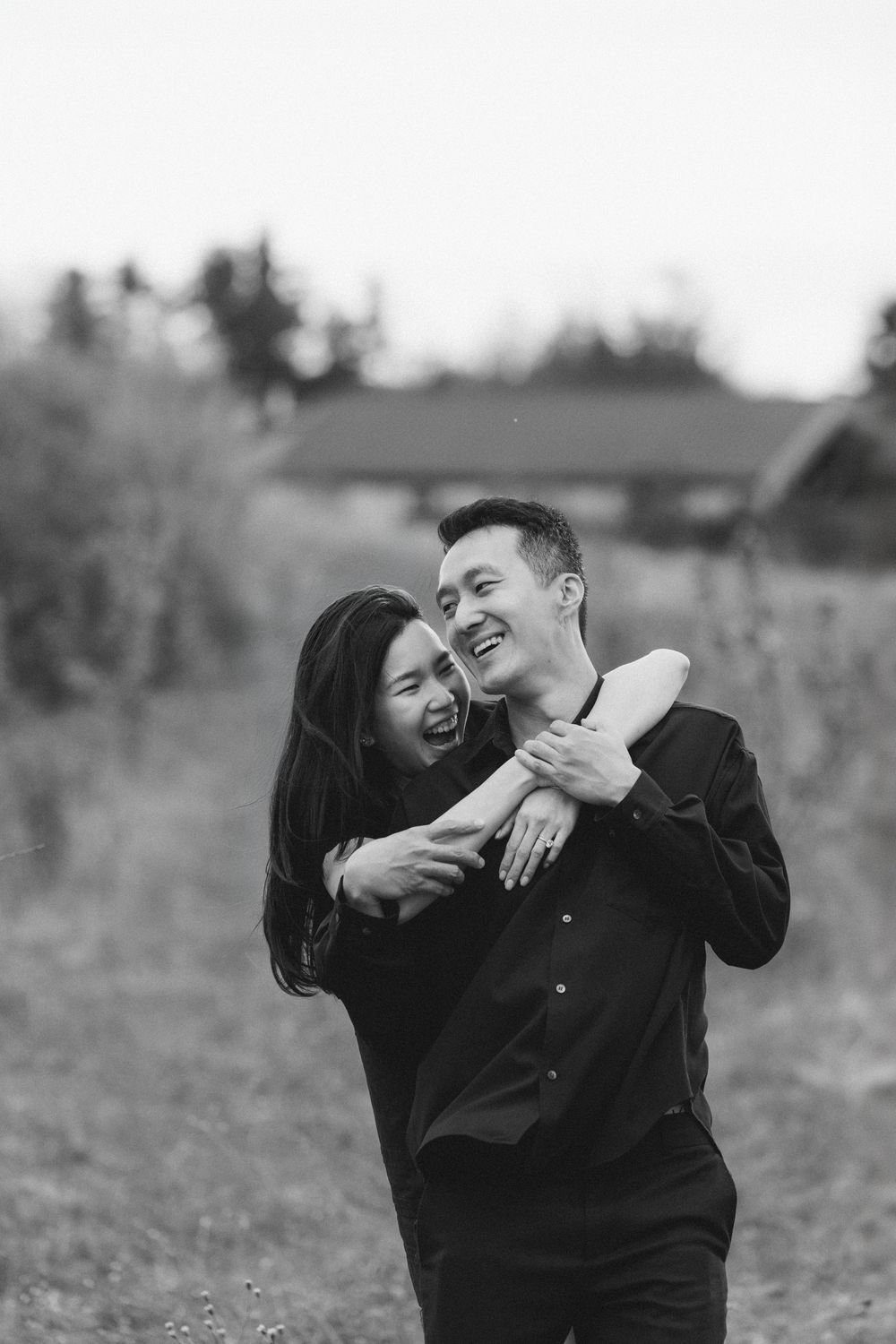 Black and white photo of a joyful couple embracing outdoors, woman hugging man from behind as he laughs.