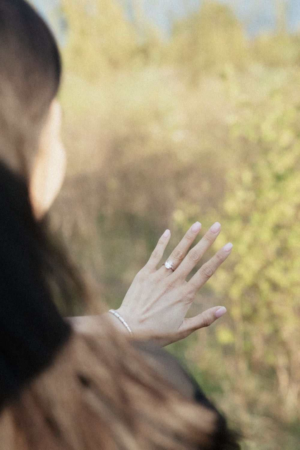 Woman with dark hair raising her hand showing an engagement ring, with blurred autumn foliage in background.