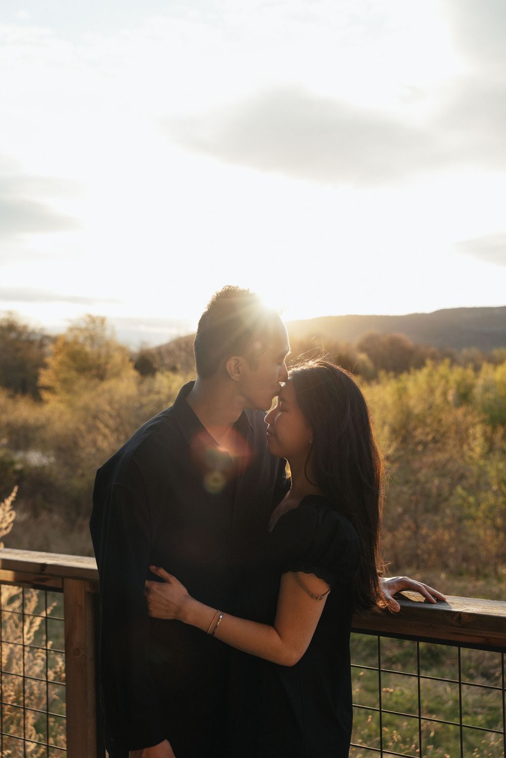 Romantic couple sharing a kiss on a deck at golden hour with scenic landscape behind them.