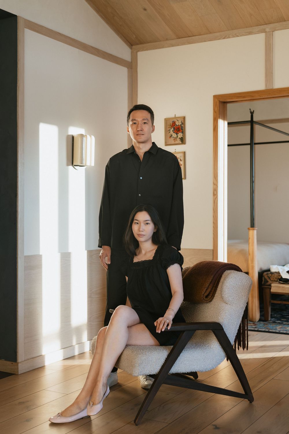 A stylish Asian couple in matching black outfits pose in a modern home interior with wood floors and natural light.
