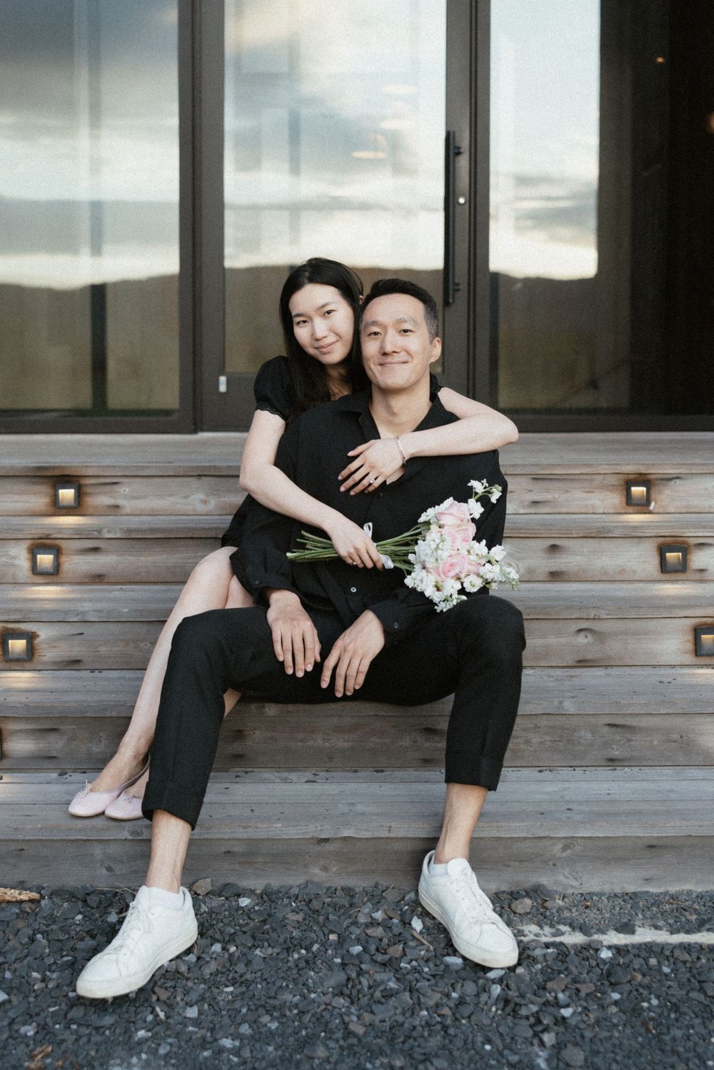 Couple in black outfits sitting on wooden steps, woman embracing man holding pink flowers.