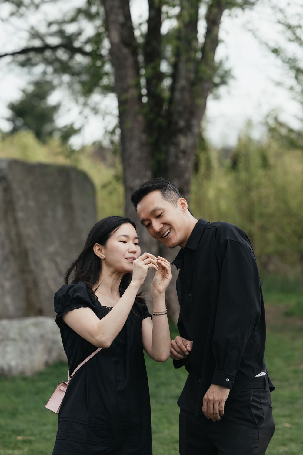A happy Asian couple in black outfits sharing a snack outdoors in a lush green park setting.