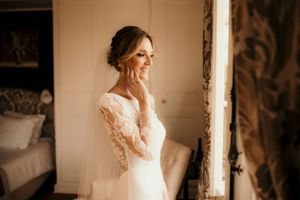 Bride in lace dress poses by window in soft natural light.
