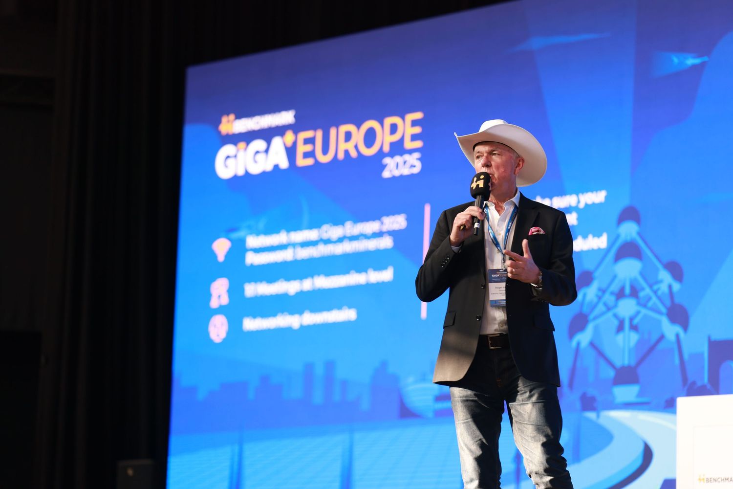 Speaker in cowboy hat presenting at Giga Europe 2025 conference on stage with blue branded backdrop.