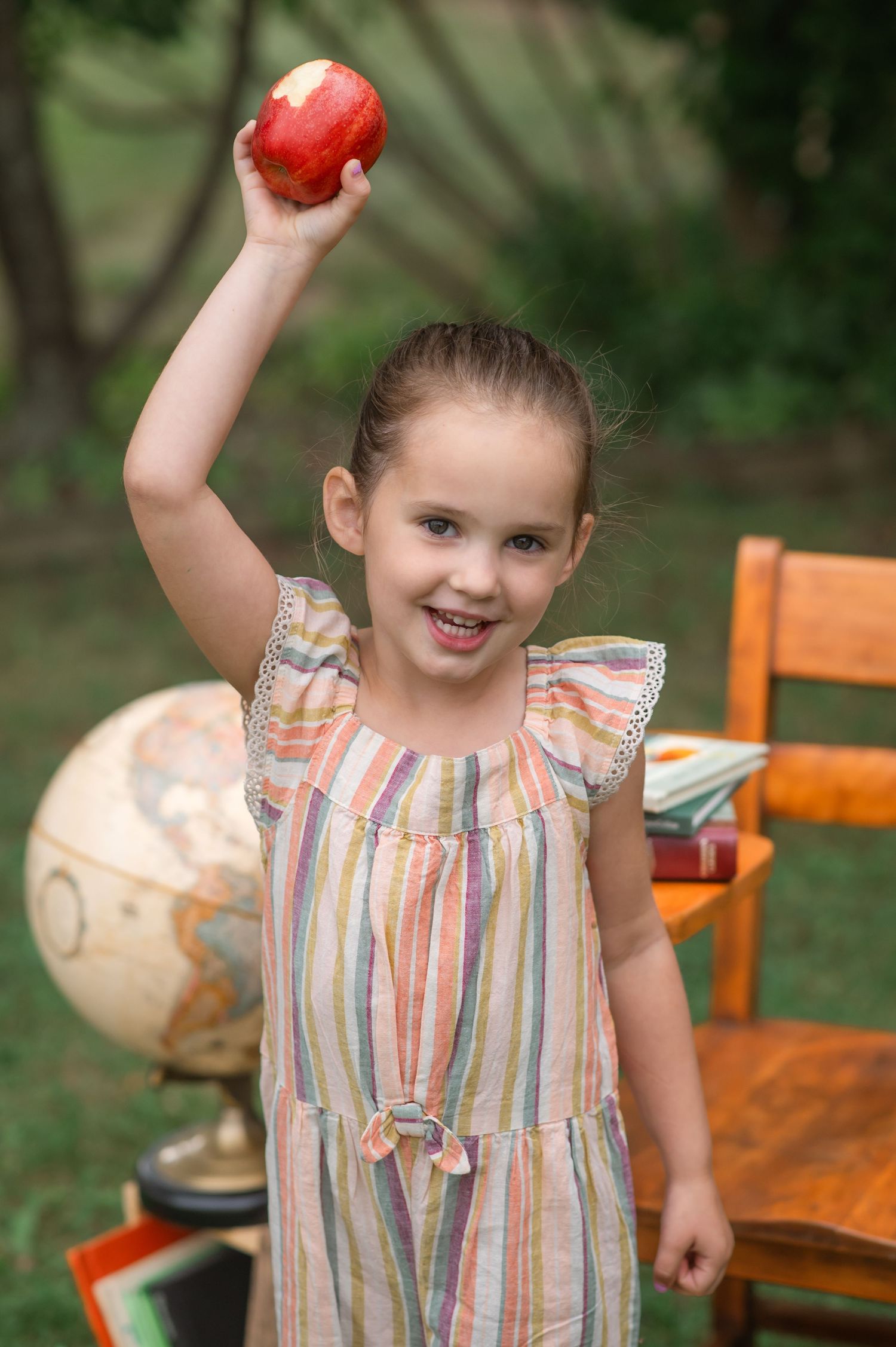 Young girl in striped dress holding a red apple up high, with a globe and books in the background.
