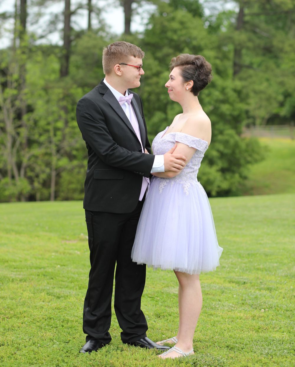 Couple in formal attire posing outdoors on green grass, woman in lavender tulle dress, man in black suit.
