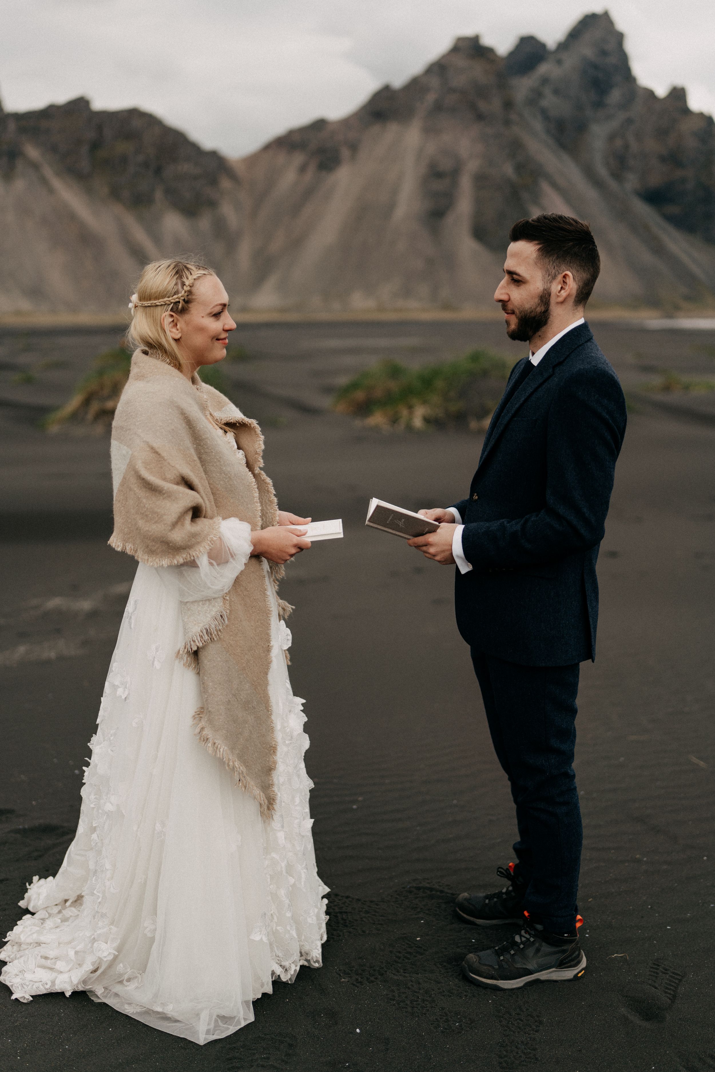 Couple exchanging vows in Vestrahorn in Iceland with dramatic mountain backdrop, bride in white gown with tan shawl.