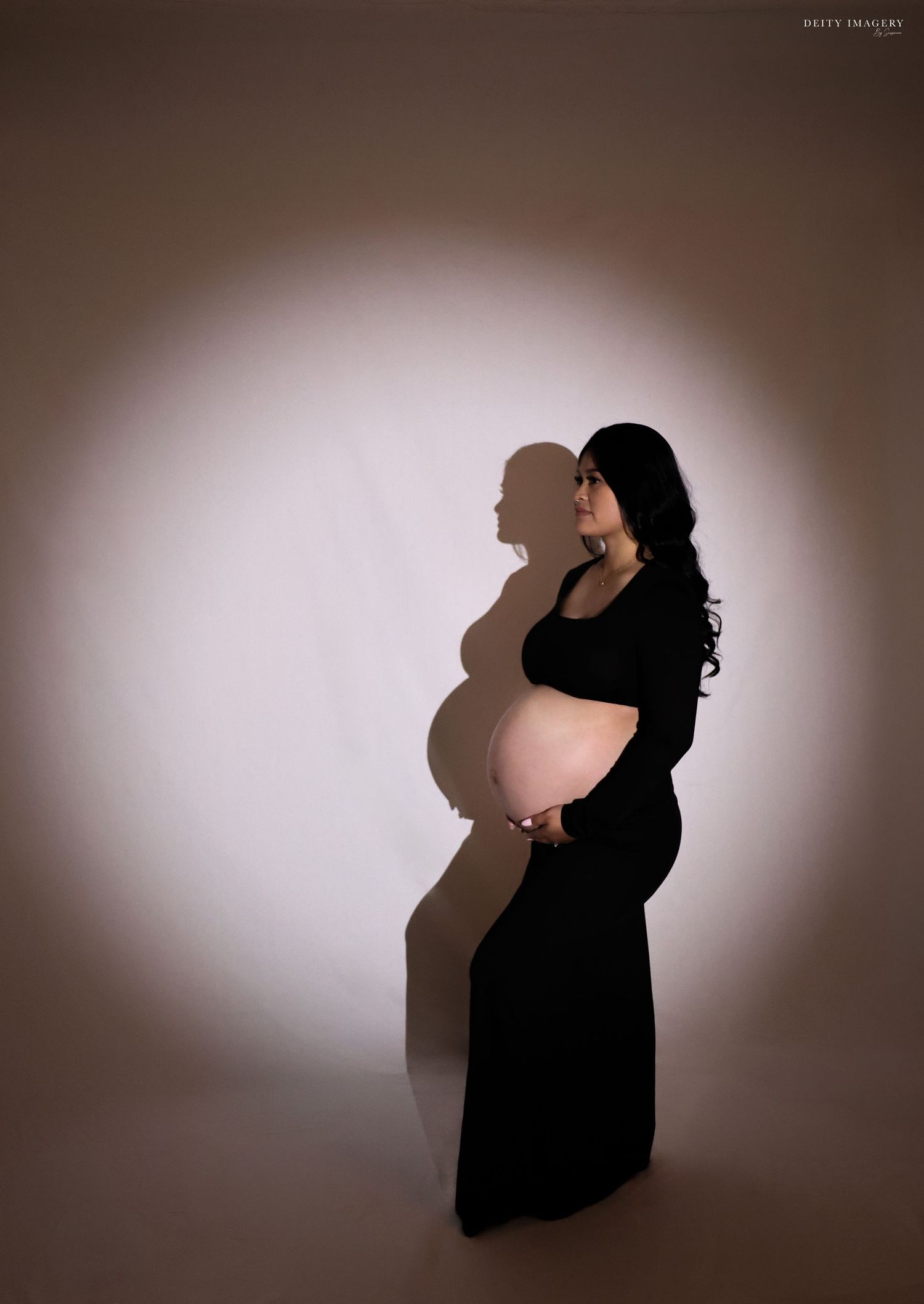 Pregnant woman in black dress holding baby bump during professional maternity photo session with dramatic lighting.