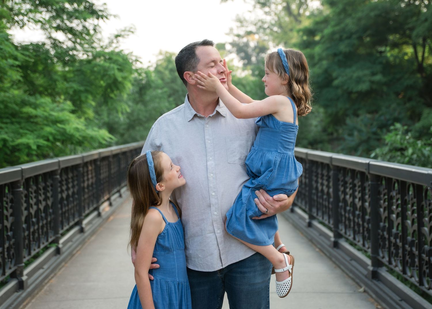 A father holds his daughter on a bridge while another daughter stands beside them, all wearing blue outfits outdoors.