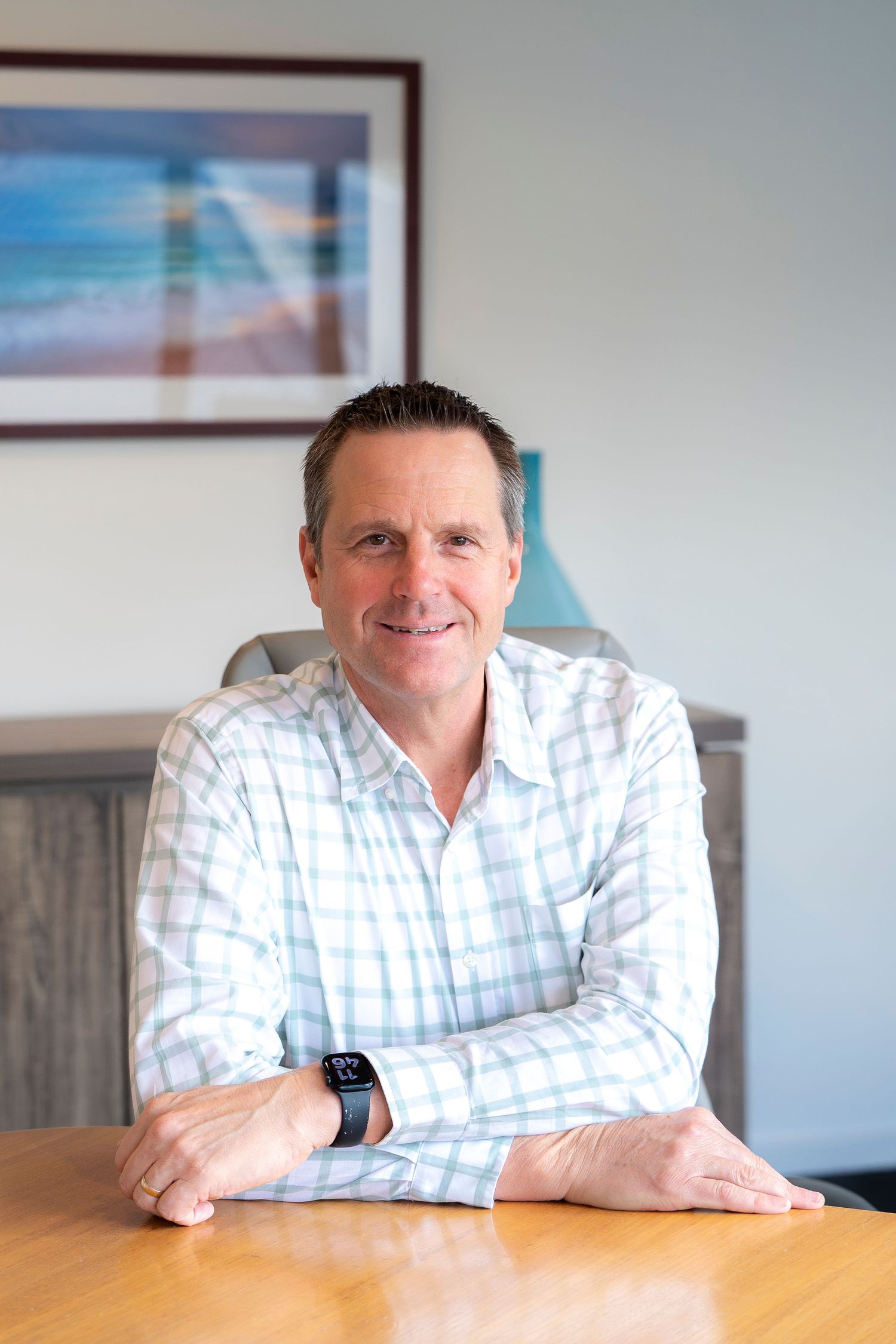 Smiling middle-aged man in plaid shirt sitting at wooden desk in modern office with coastal artwork.