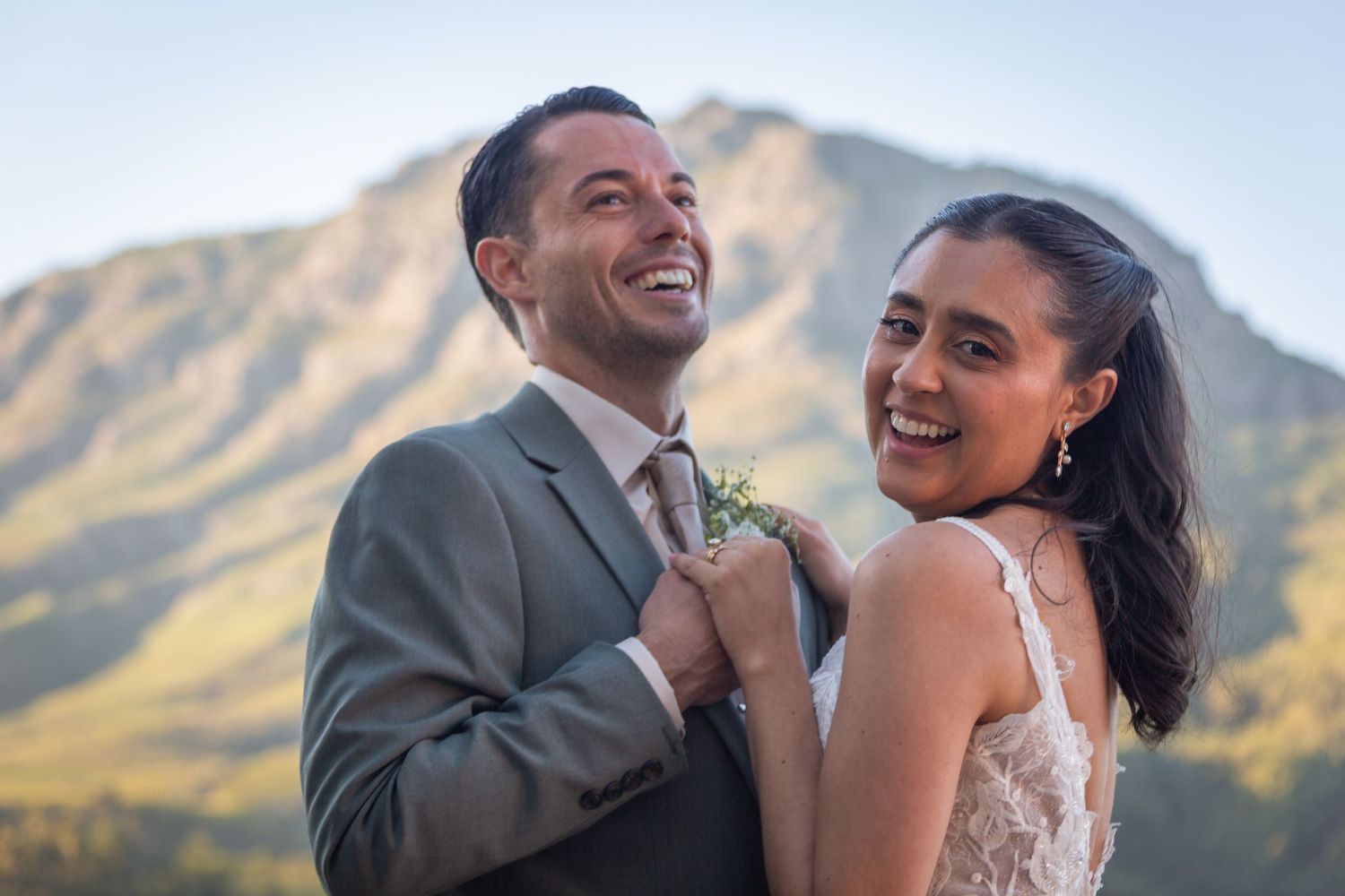 Happy couple in wedding attire laughing together with a scenic mountain backdrop on their wedding day.