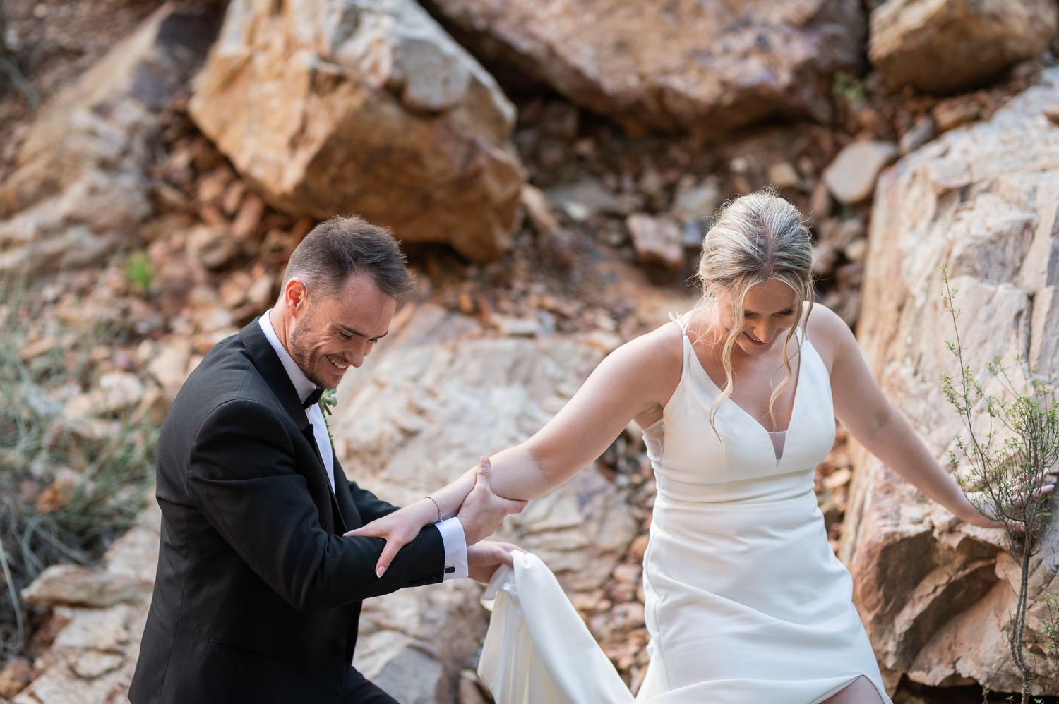 Groom in black tuxedo helps bride in white gown navigate rocky terrain during outdoor wedding ceremony.