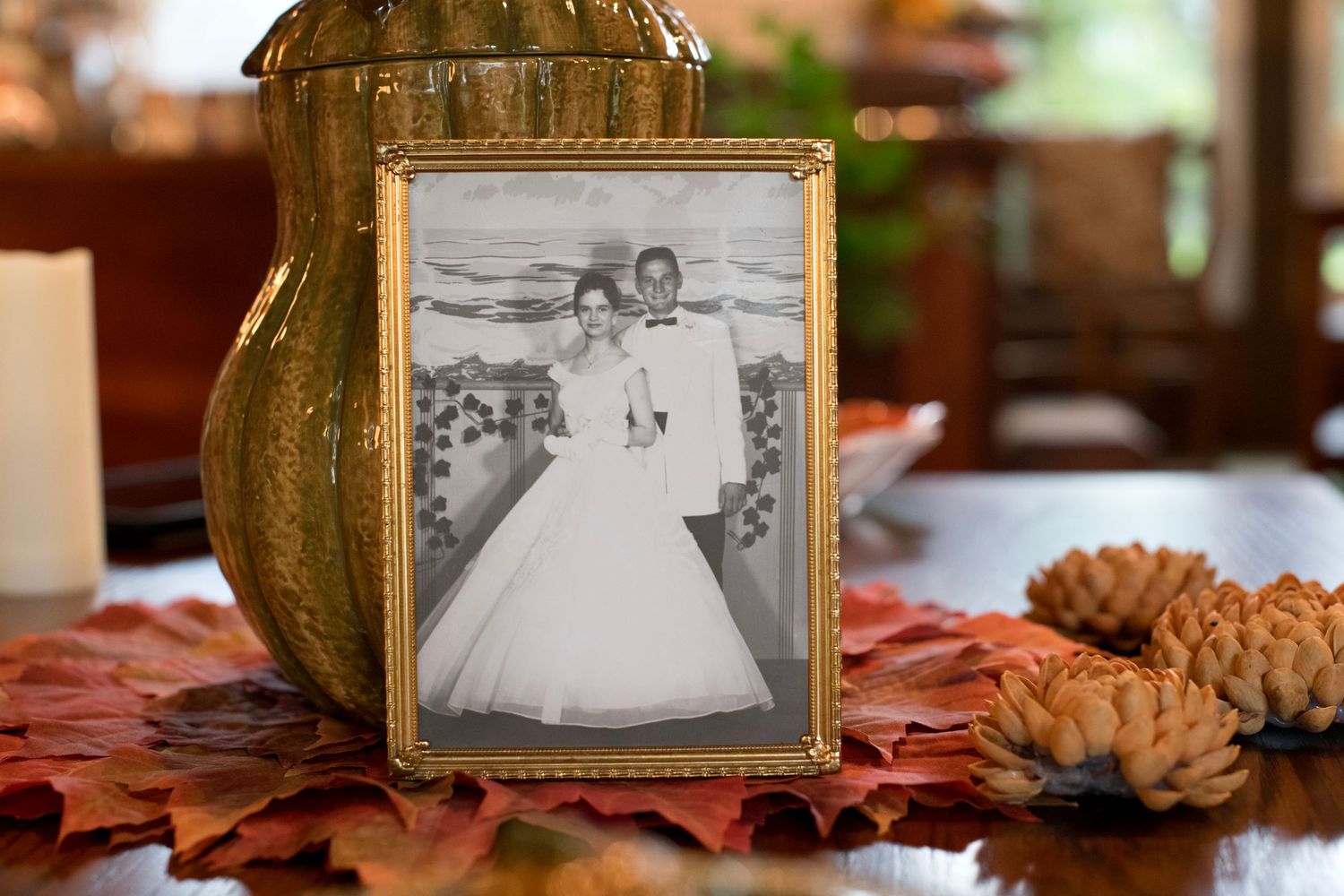 Vintage black and white wedding photo in gold frame surrounded by autumn leaves and pine cones on wooden table.