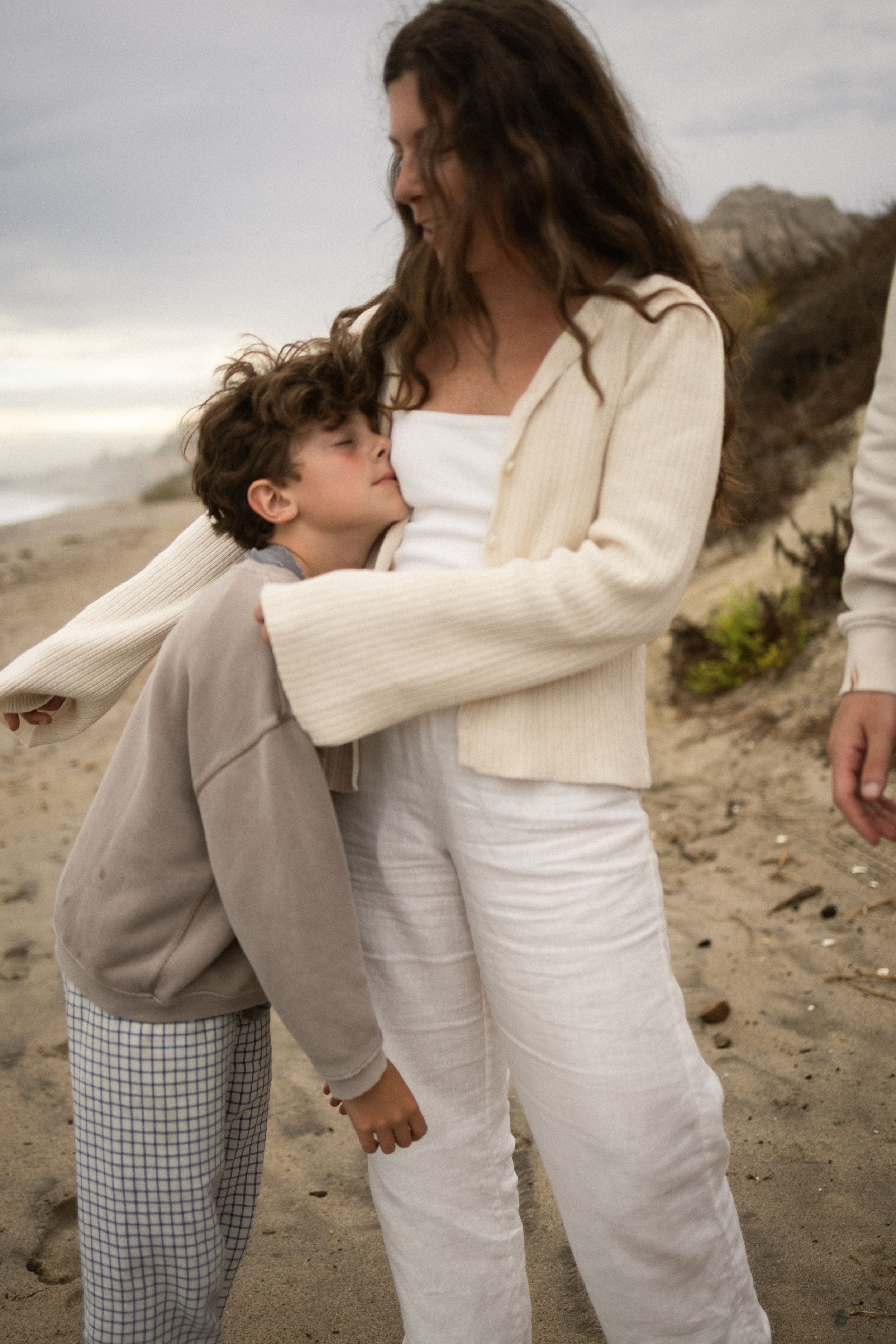 A young boy hugs his smiling mother on a sandy beach, both wearing neutral tones.
