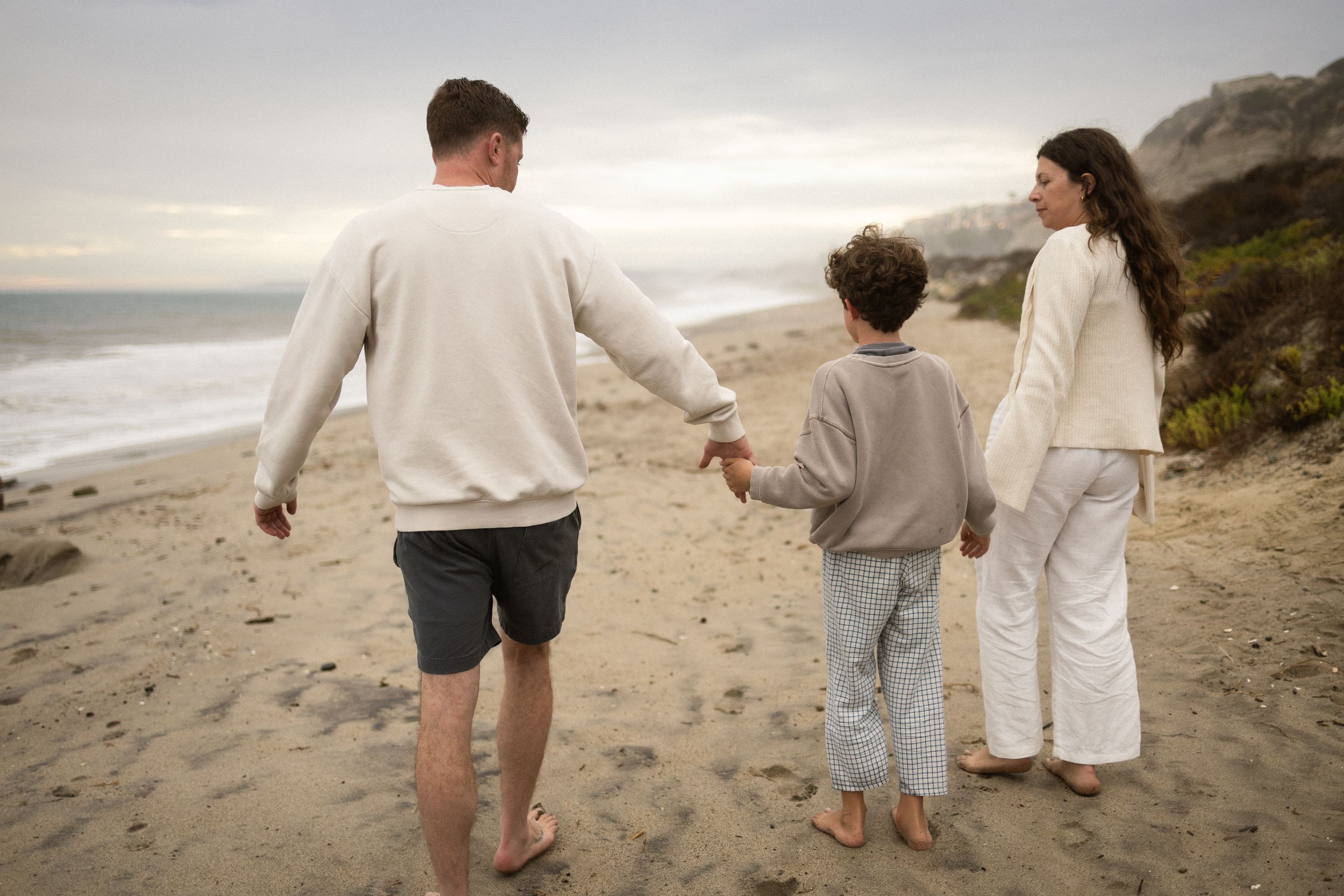 Family of three walking hand-in-hand on a sandy beach at dusk, wearing casual neutral-toned clothing.