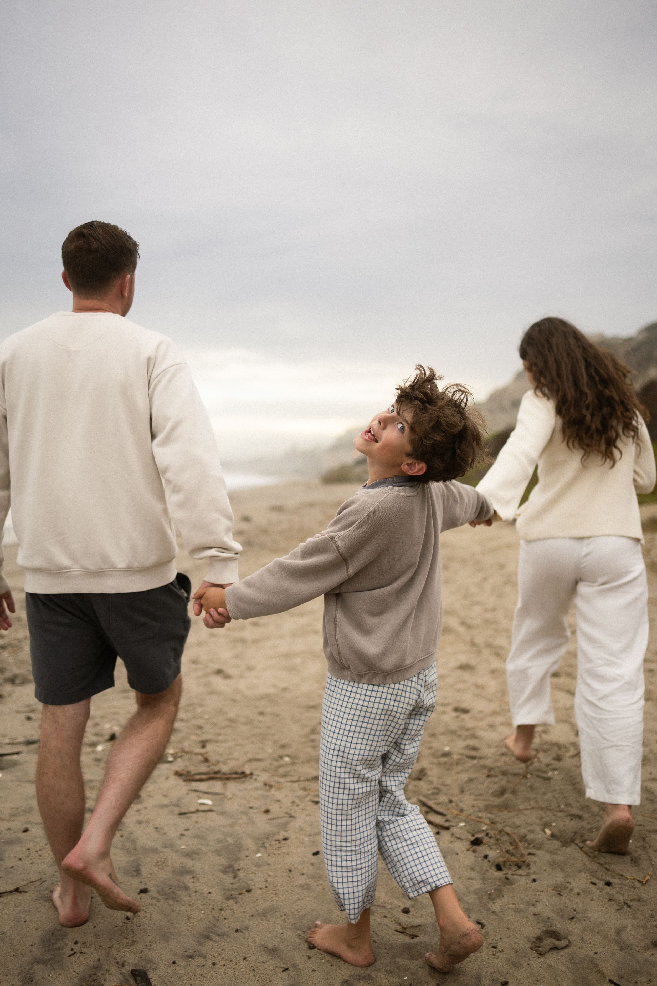 Family walking barefoot on sandy beach, child looking up at parents while holding hands on cloudy day.