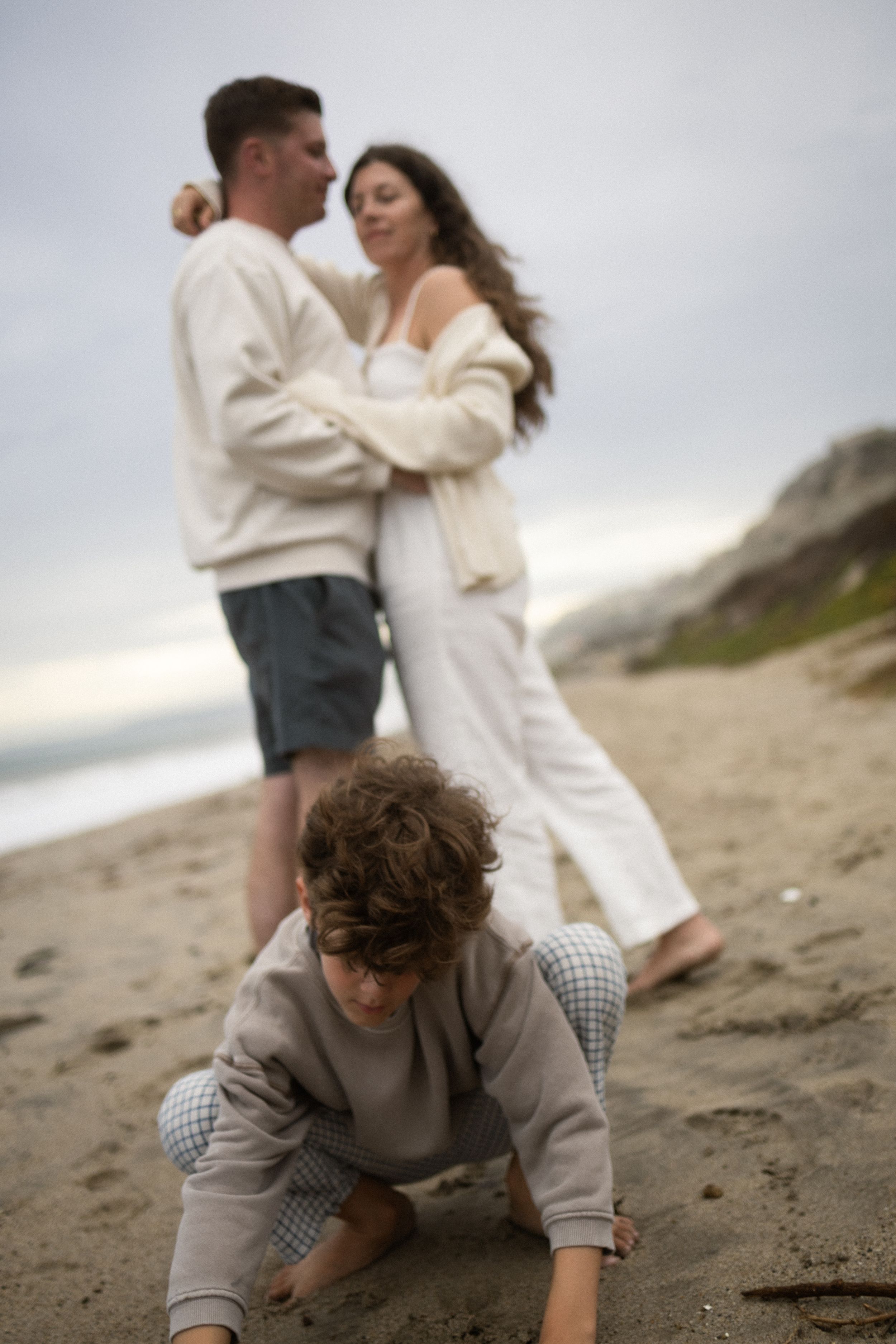 Couple embracing on beach while young curly-haired child plays in sand in foreground.