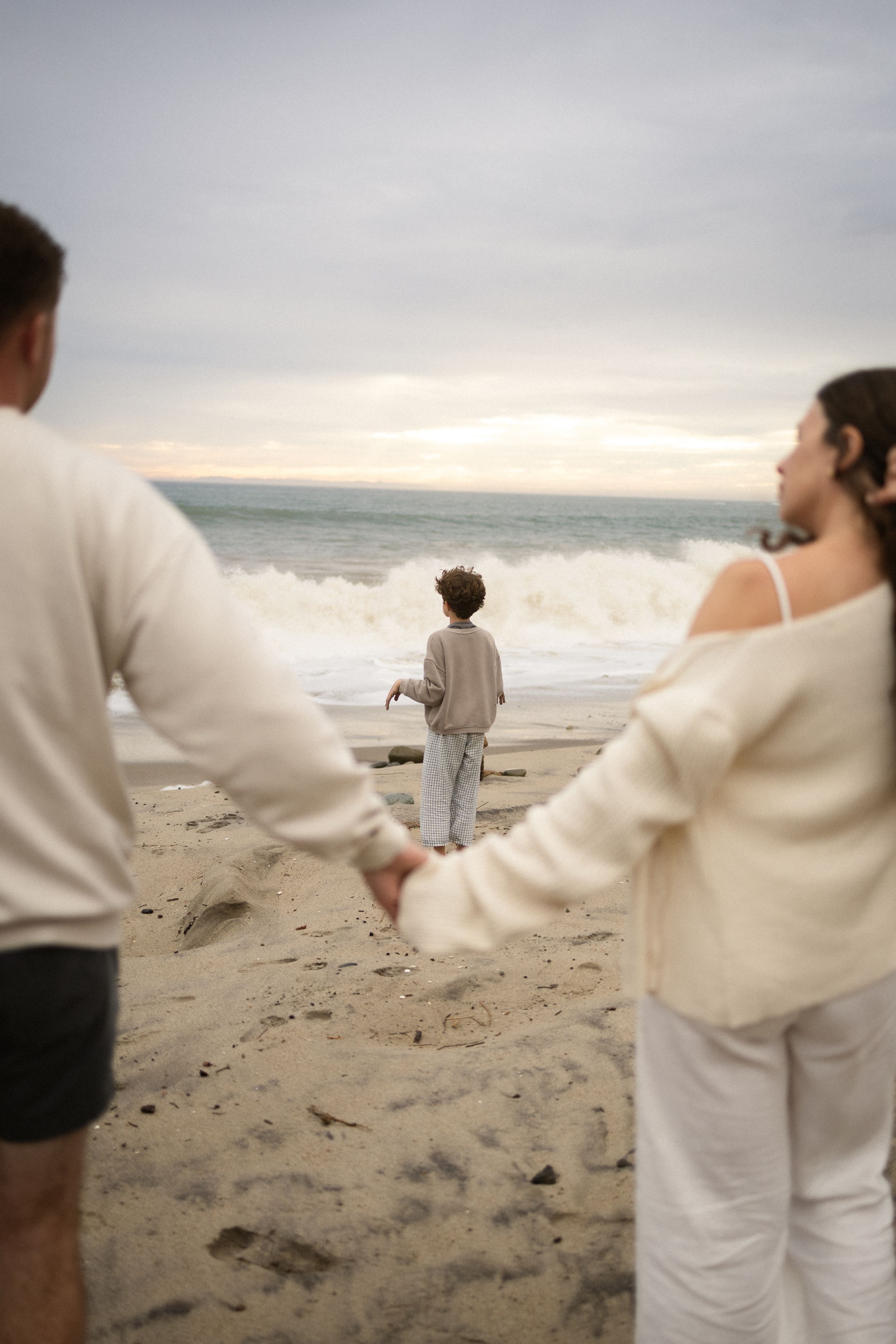 A family holds hands on a sandy beach while a child stands at the ocean's edge watching waves under a cloudy sky.