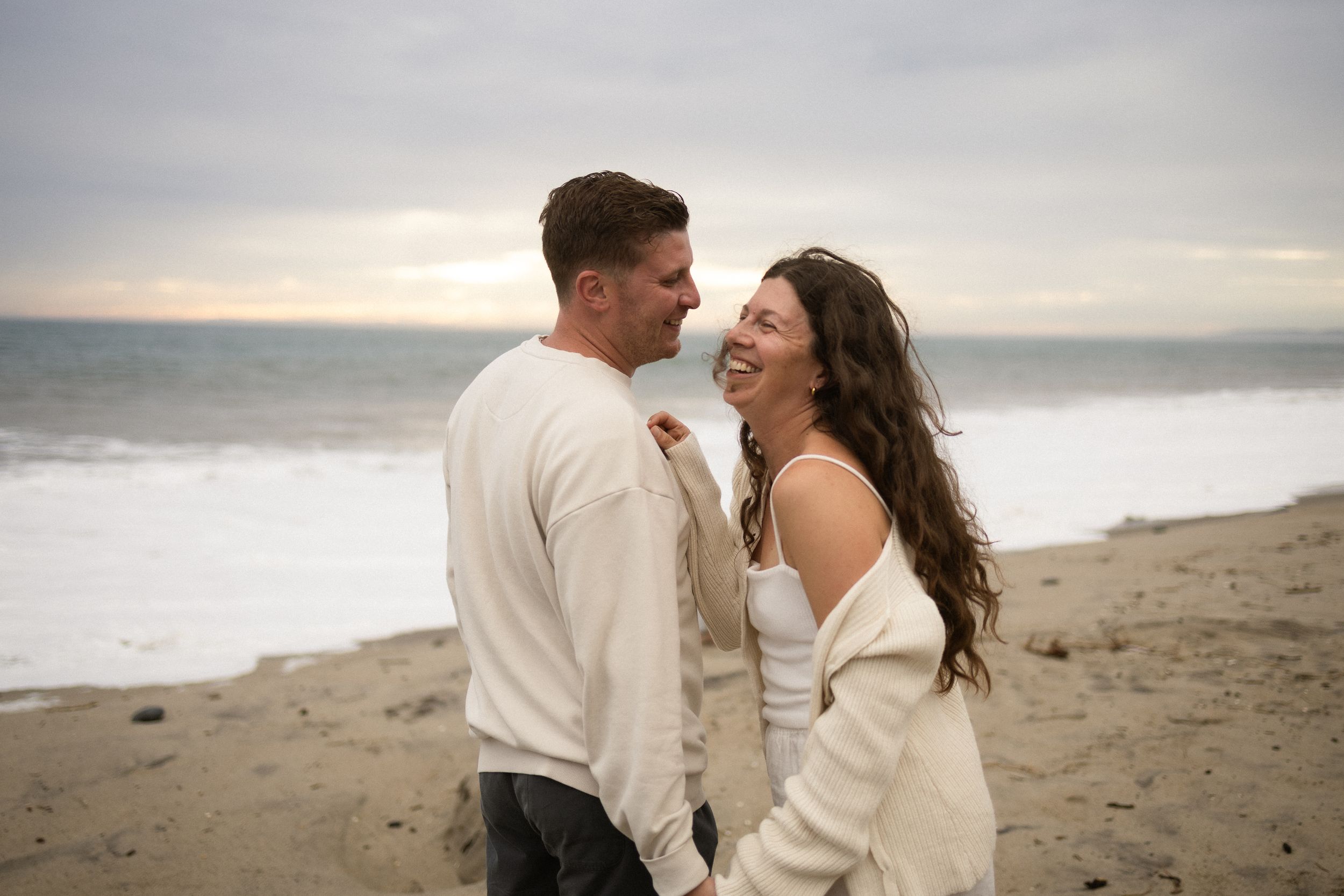 A smiling couple in cozy white sweaters shares a romantic moment on a sandy beach with ocean waves at sunset.