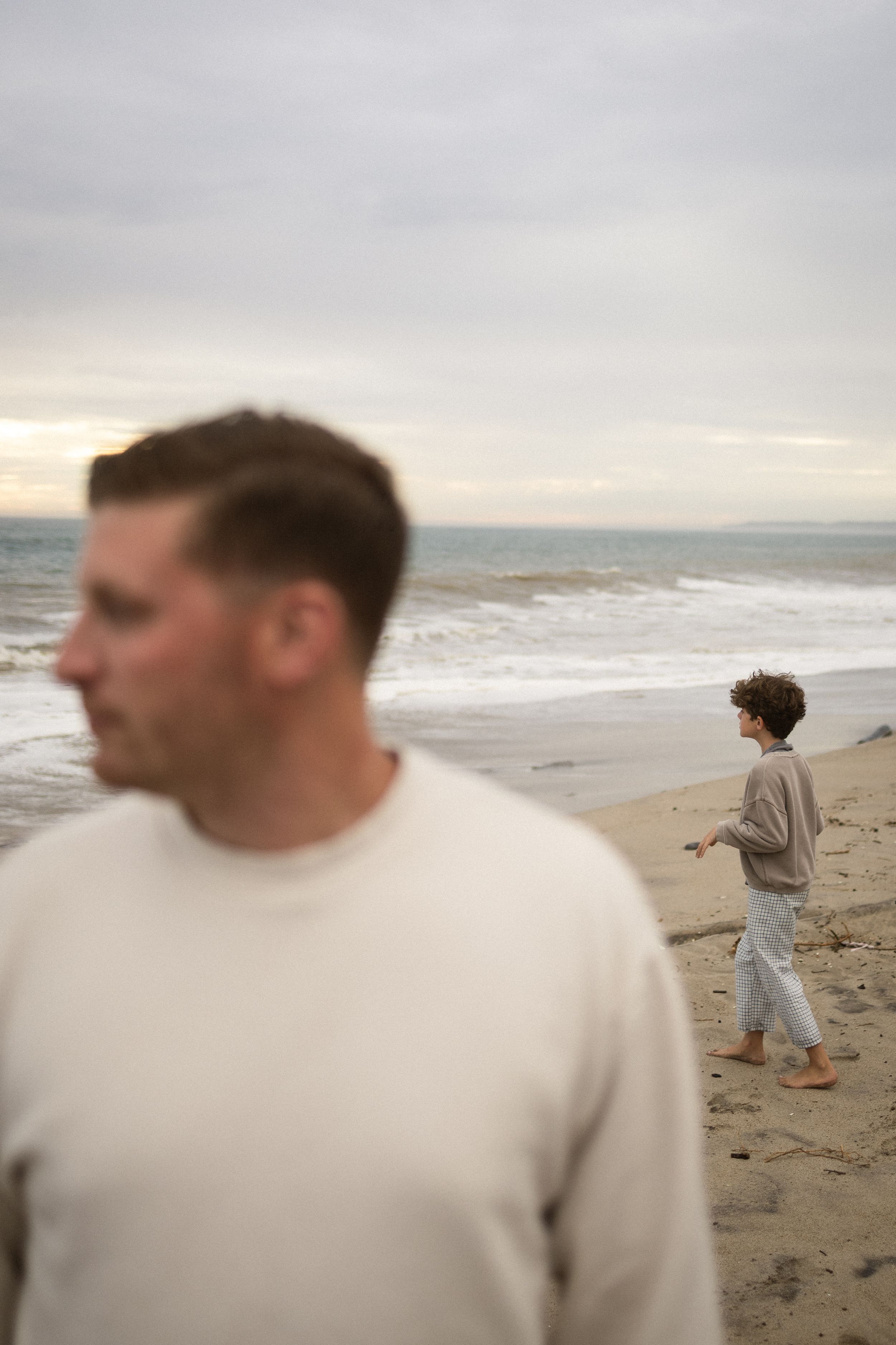 Man in white sweater stands on beach while child plays near ocean waves under cloudy sky.