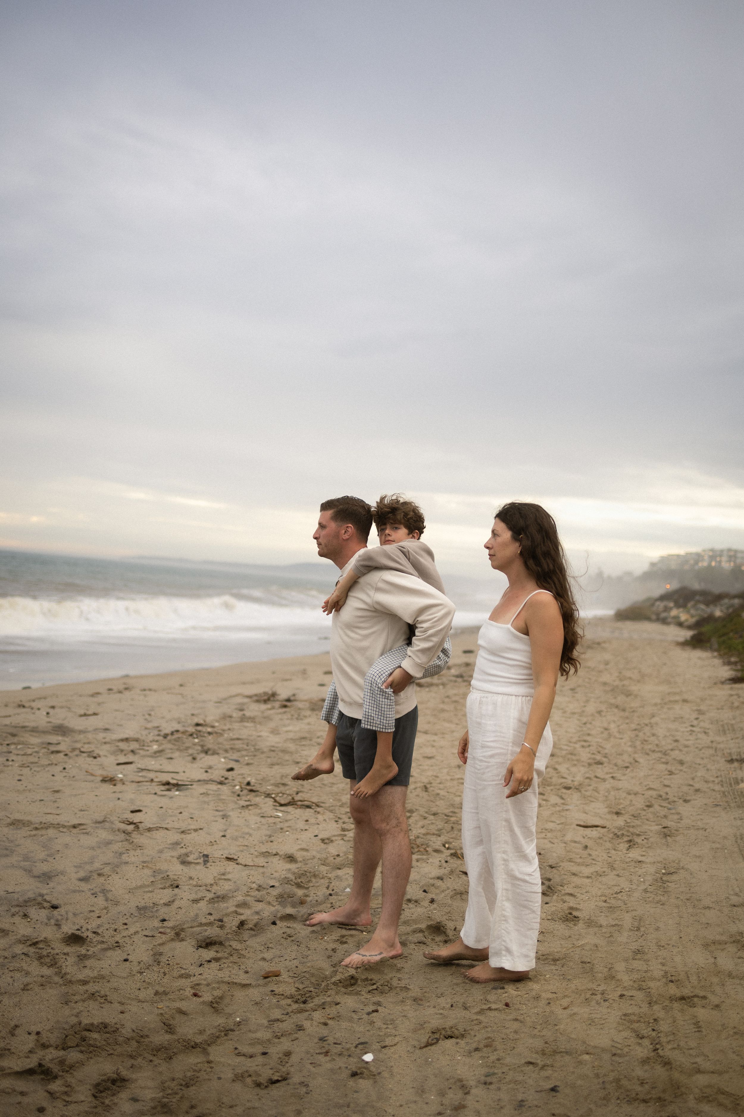 Family of three standing barefoot on a sandy beach, gazing at ocean waves under a cloudy sky.
