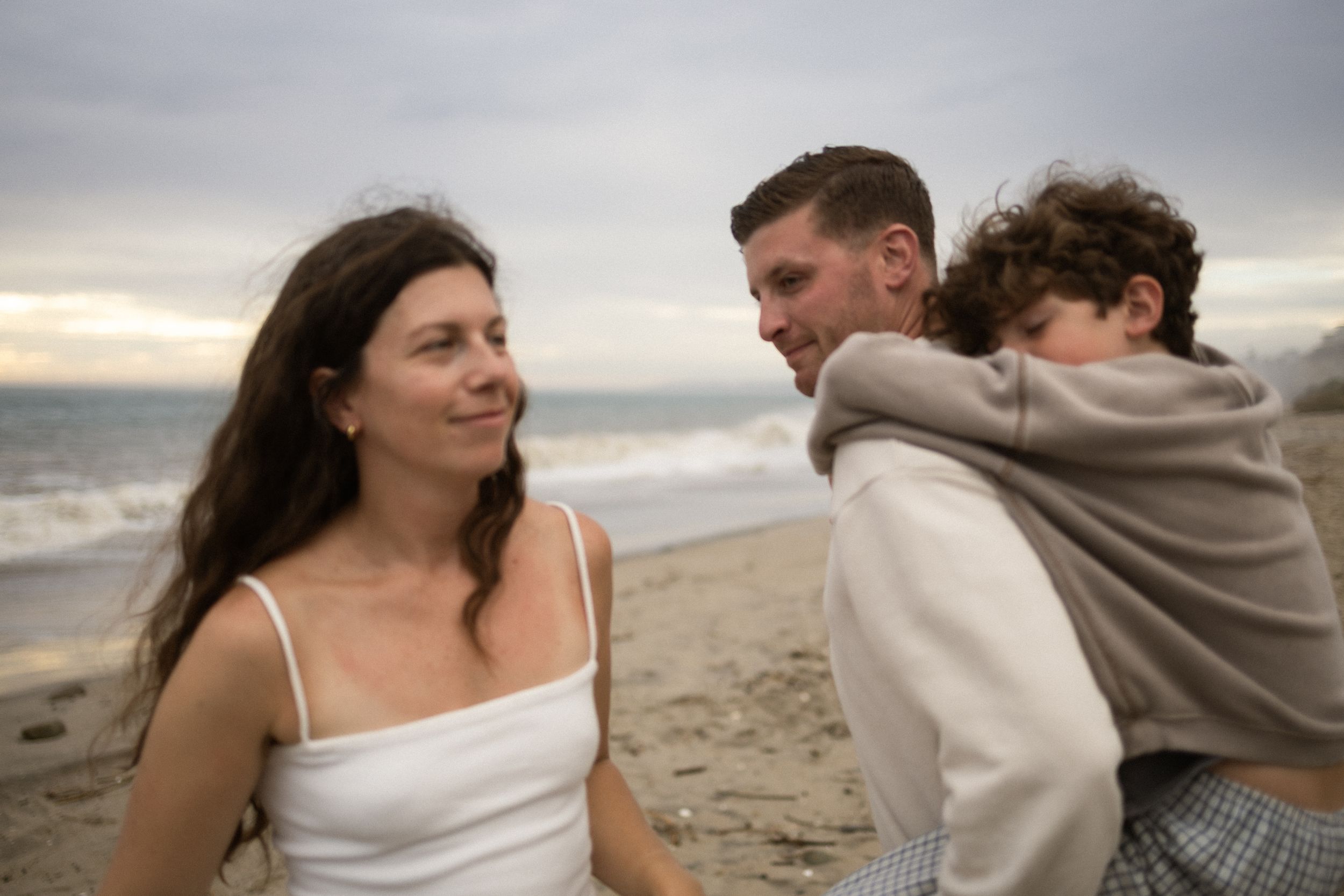 Family of three sharing a tender moment on a sandy beach at sunset, with ocean waves in the background.