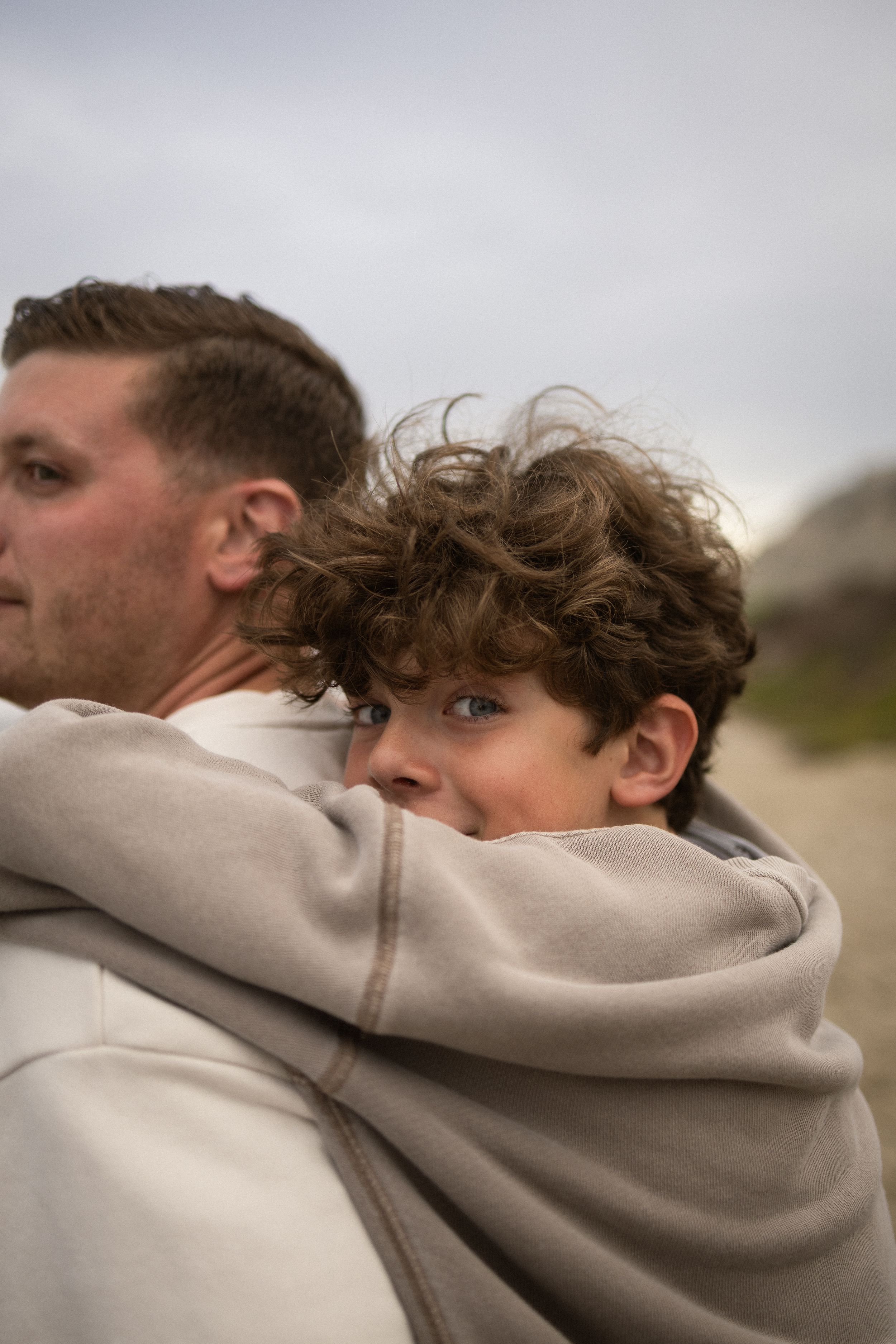 A young curly-haired boy peeks over a man's shoulder on a cloudy beach day.