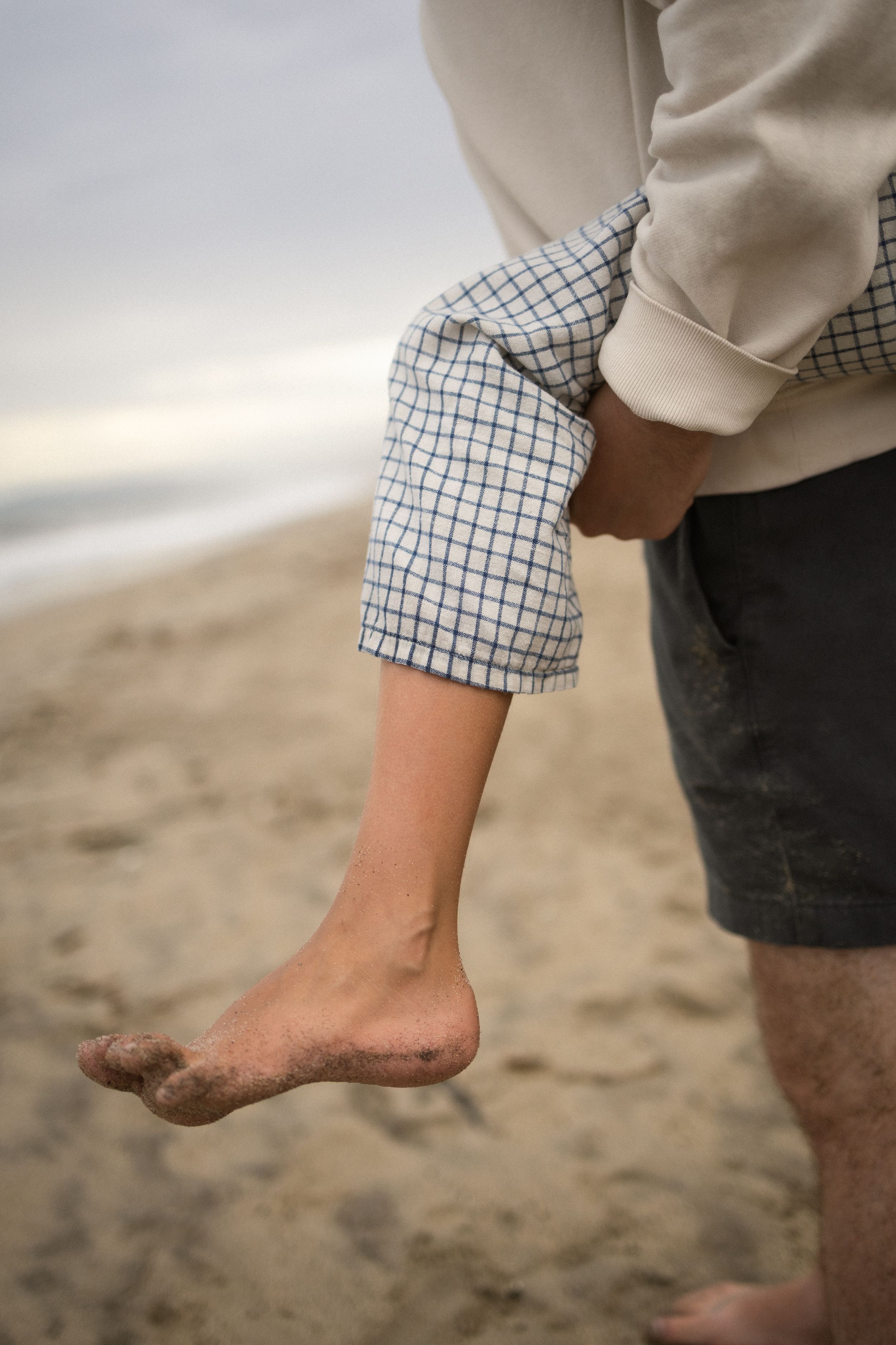 Close-up of a child's sandy bare foot dangling as an adult carries them on a beach.