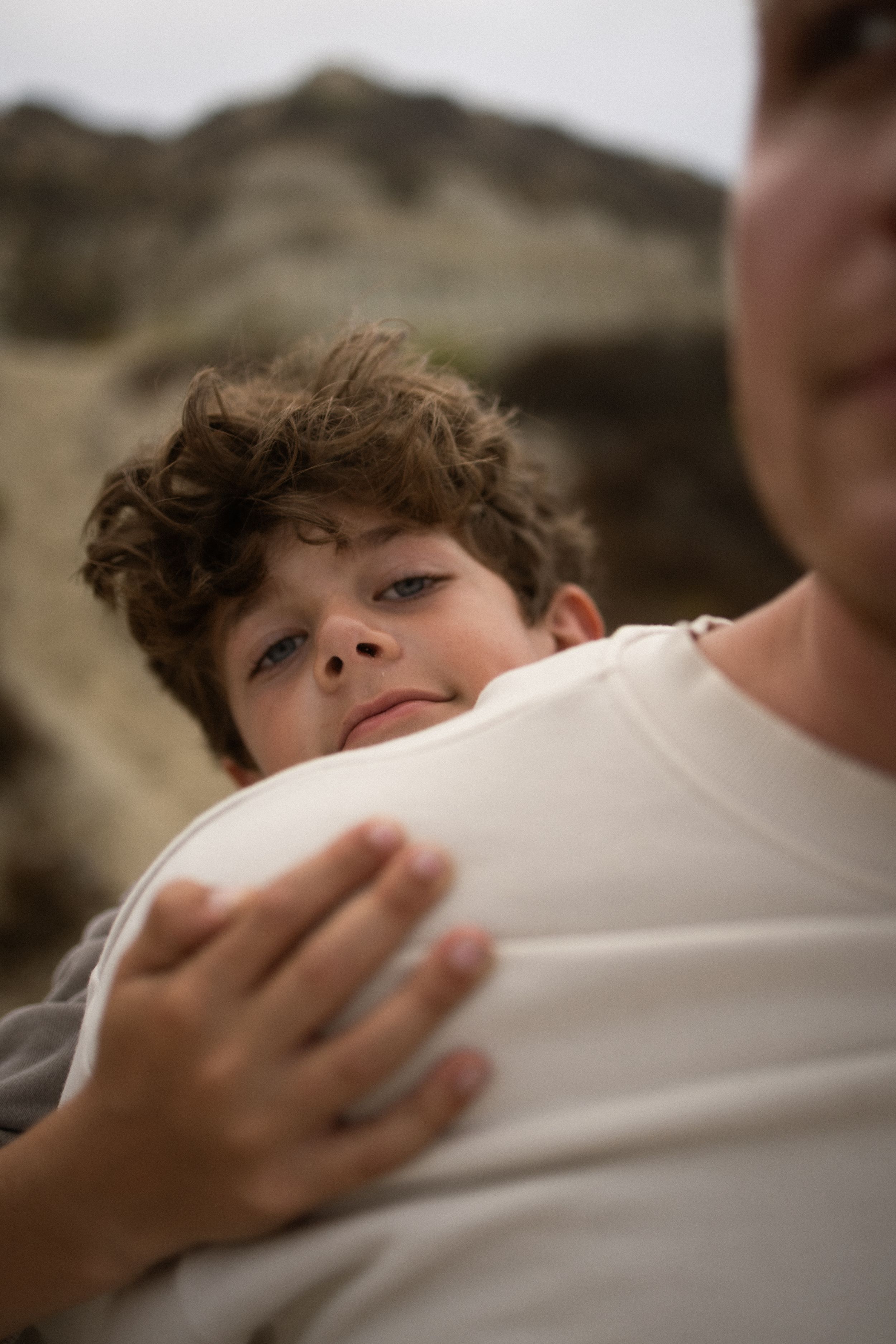A curly-haired boy smiles while hugging an adult in a white shirt outdoors near rocky terrain.