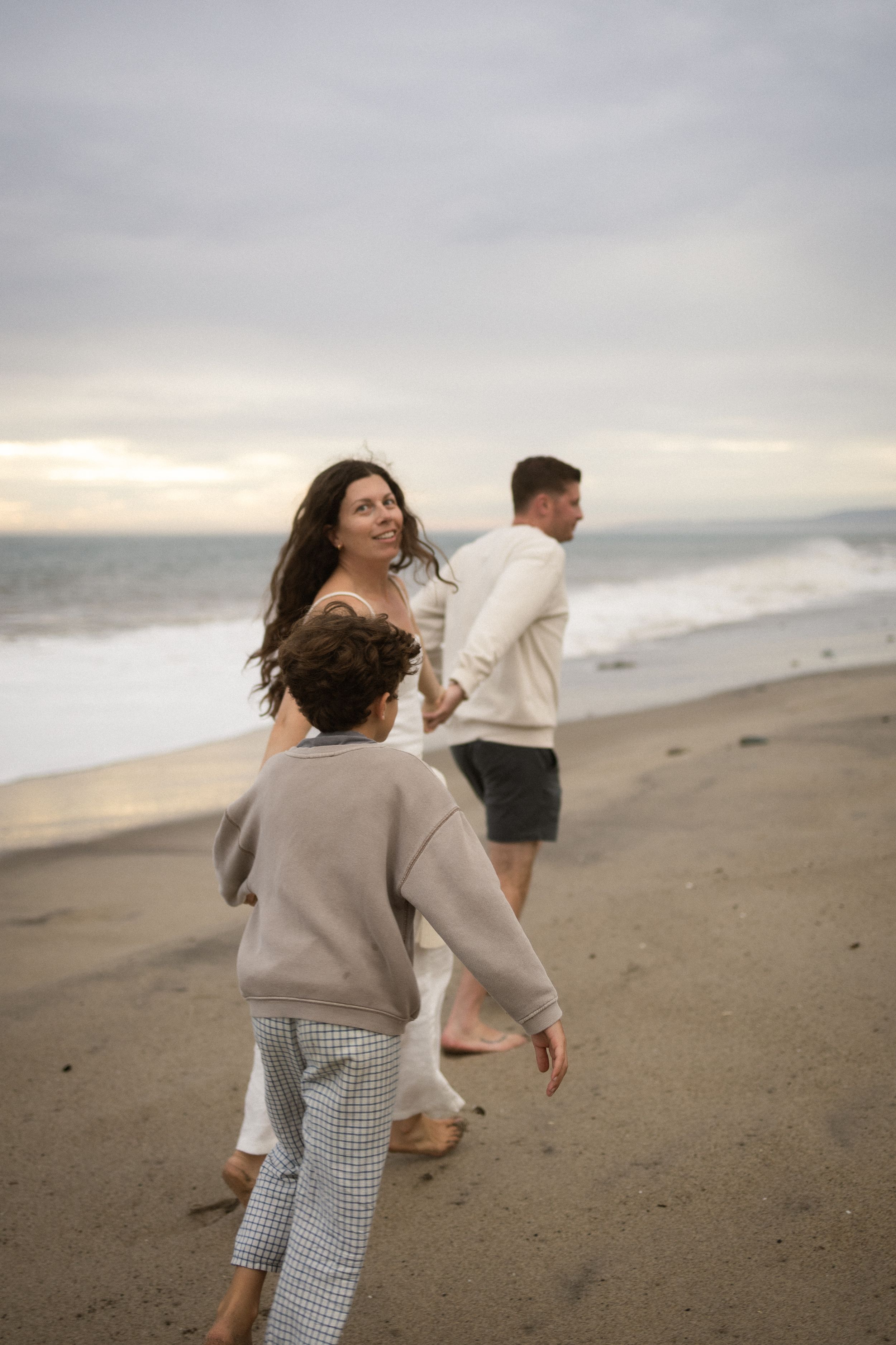 A happy family walks along a sandy beach at sunset, with a woman smiling back at her child.
