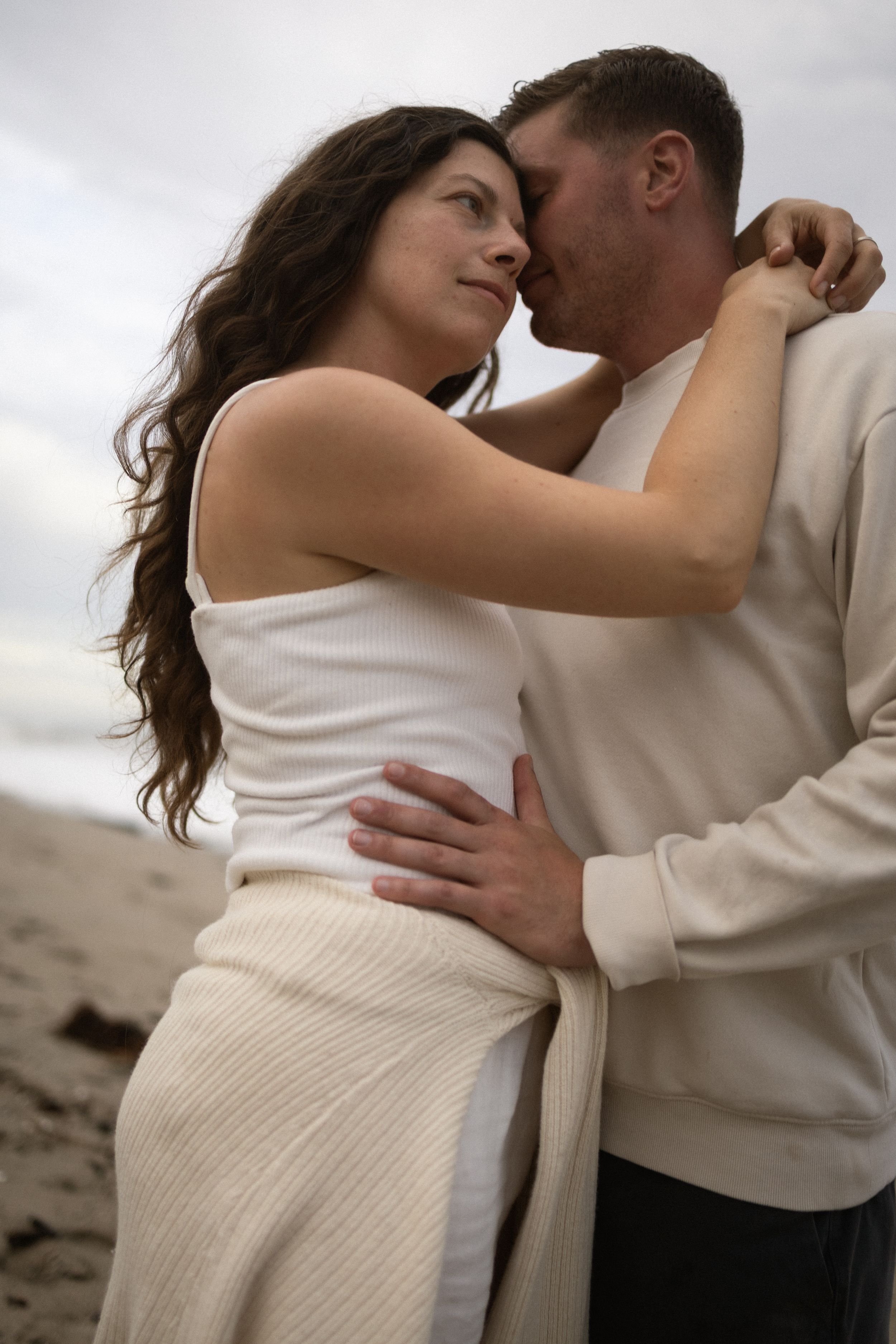 A couple embraces intimately on a beach, woman in white dress gazing up at man in cream sweater under cloudy skies.