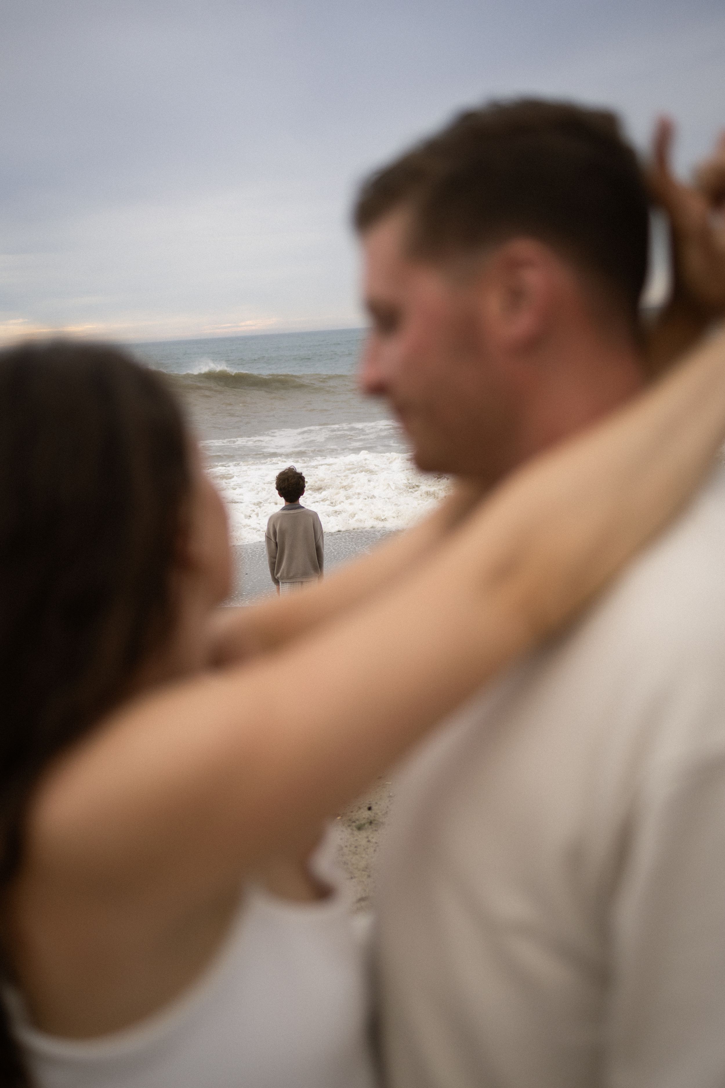 Couple embracing on beach while child stands alone at ocean's edge on cloudy day.