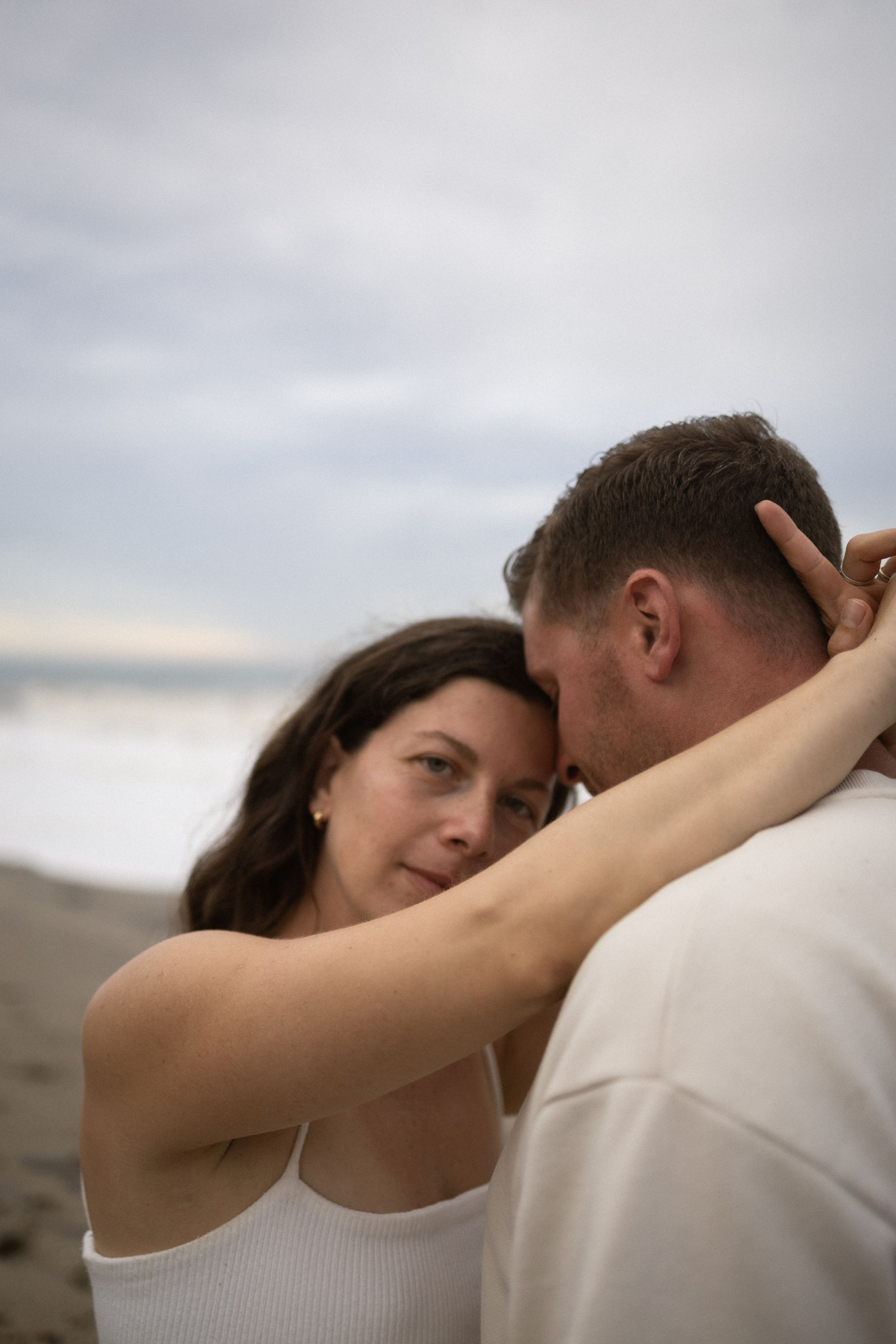 A couple embraces tenderly on a beach at dusk, the woman gazing softly while resting her hand on his neck.