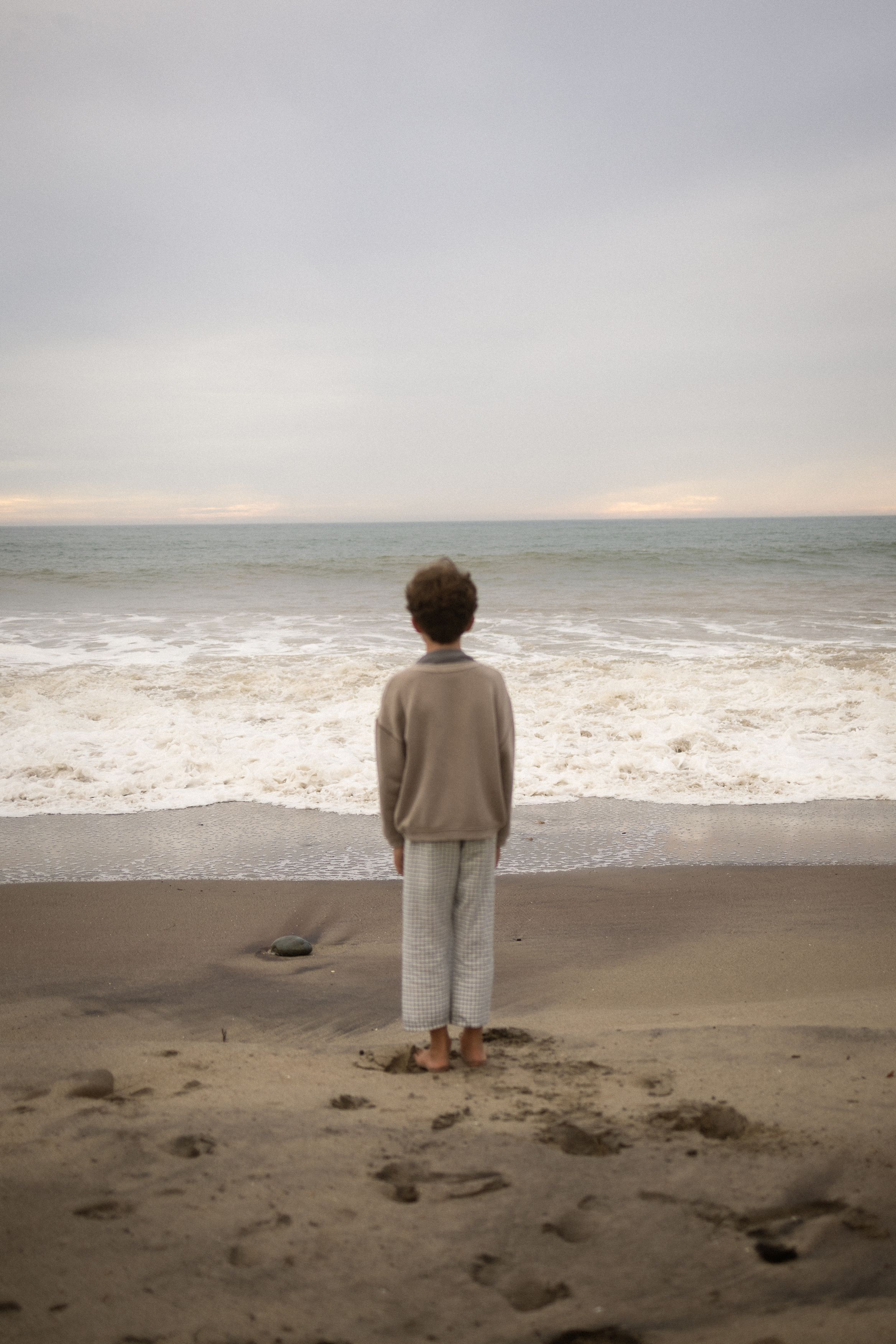 Person standing barefoot on a sandy beach, gazing at ocean waves under a cloudy sky.
