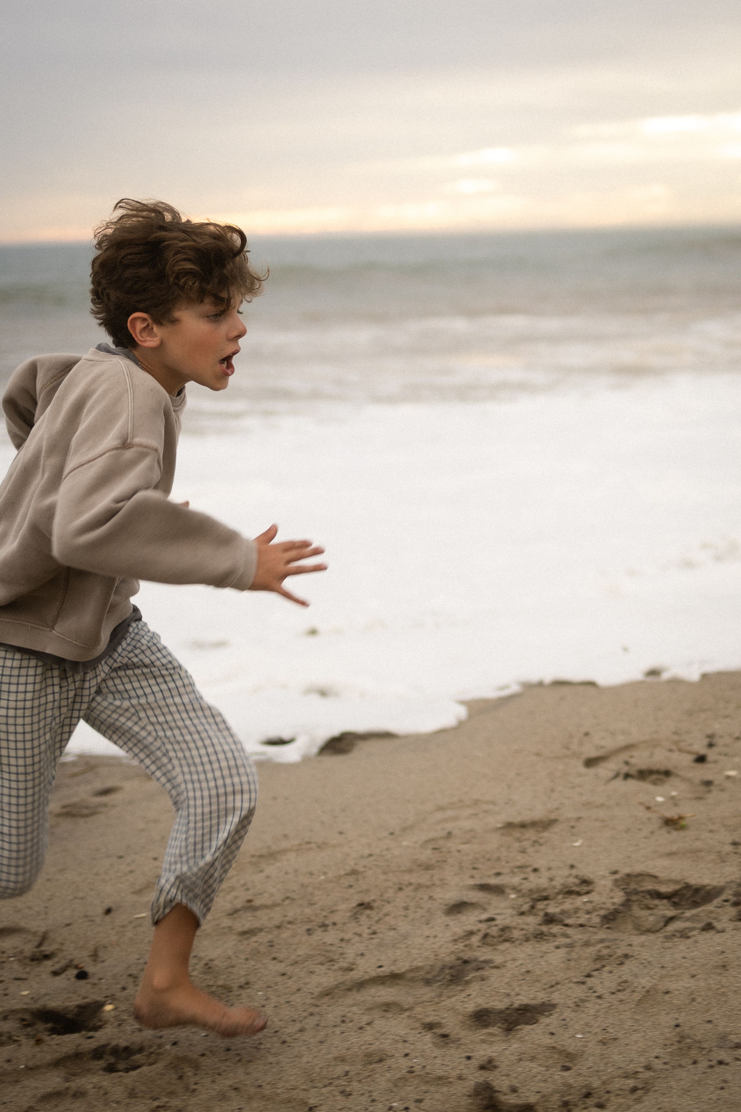 A barefoot child runs along a sandy beach at dusk, wearing a beige hoodie and plaid pants with ocean waves behind.