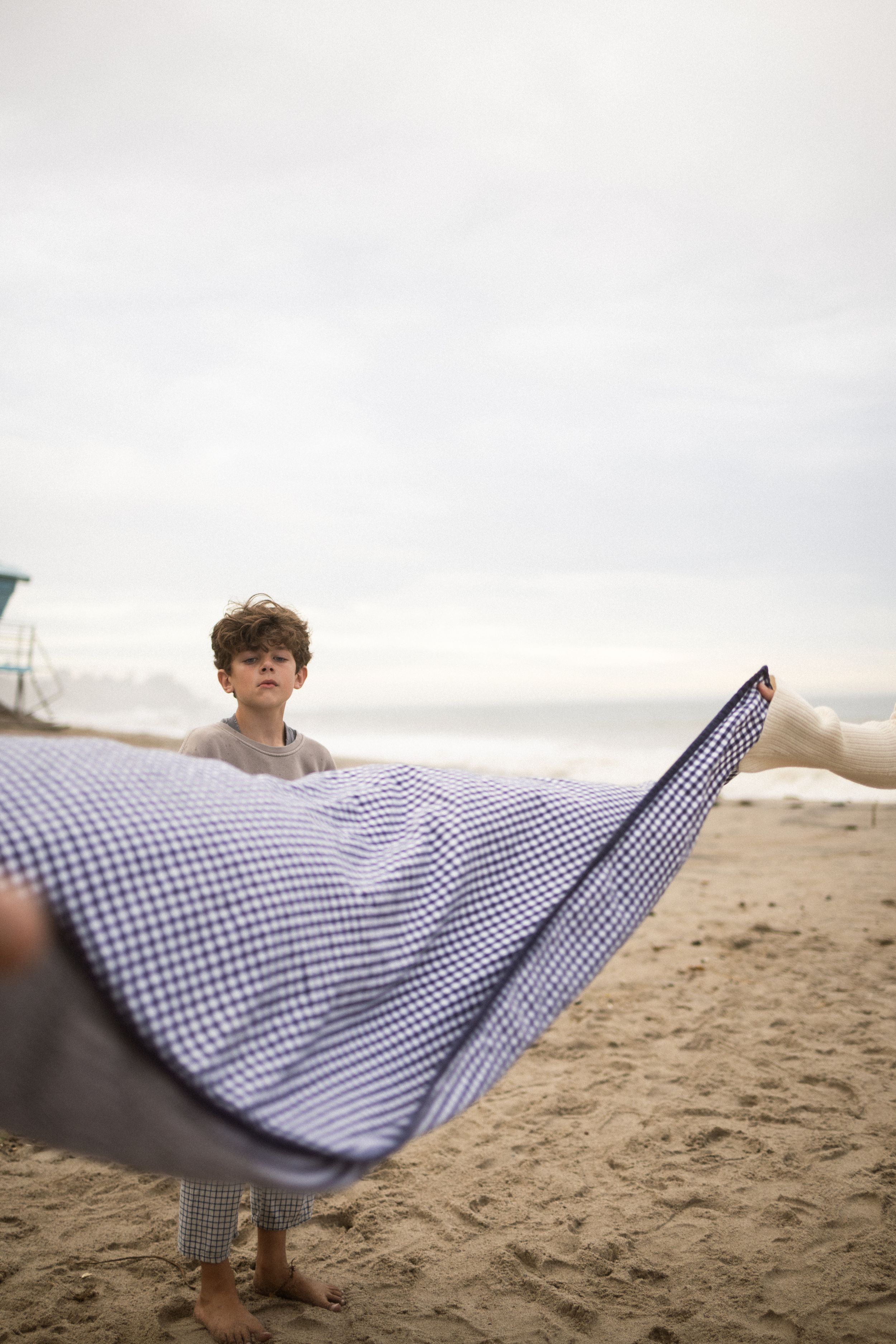 A young boy stands barefoot on a sandy beach holding a blue gingham blanket billowing in the wind.
