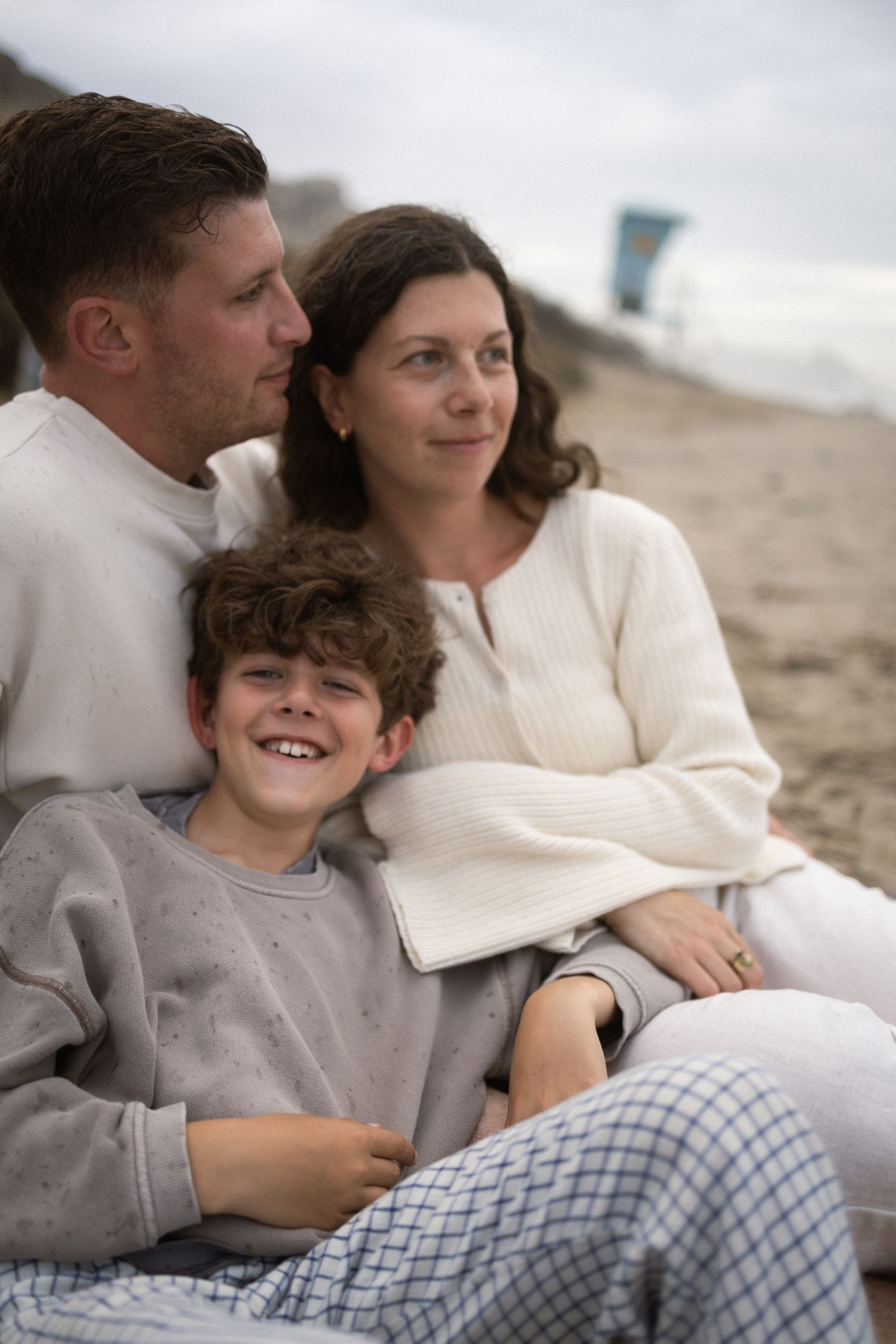 A happy family of three sits together on a beach, smiling and enjoying a cloudy day outdoors.